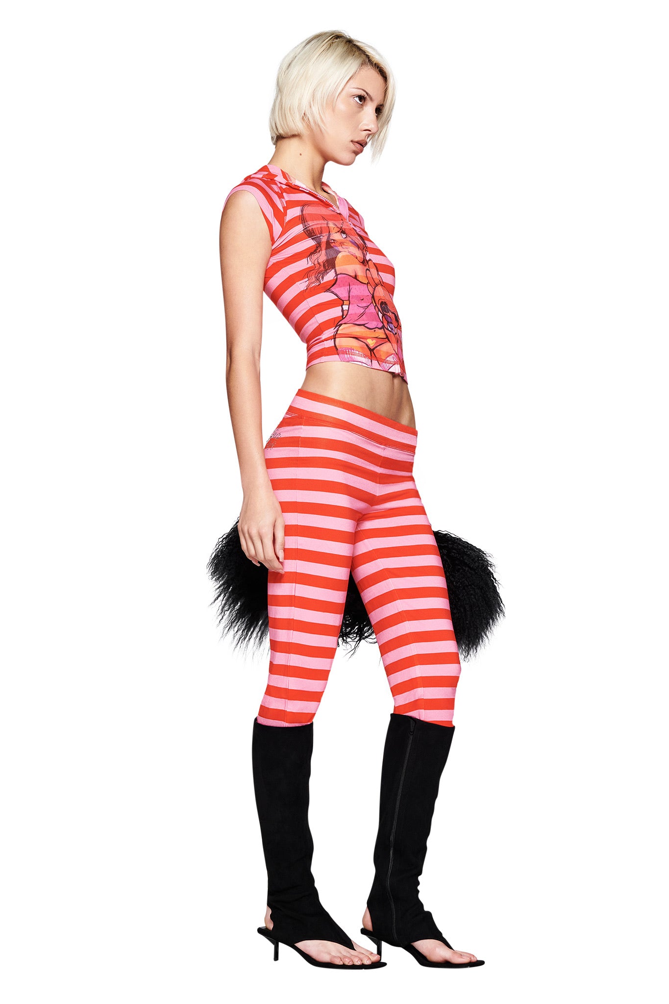 A woman with short blonde hair channels Fafinettes vibes in the I.AM.GIA FAFI BLARE CAP SLEEVE HOODIE - PINK STRIPE, styled with black open-toe stiletto boots and furry black accessories against a white background.