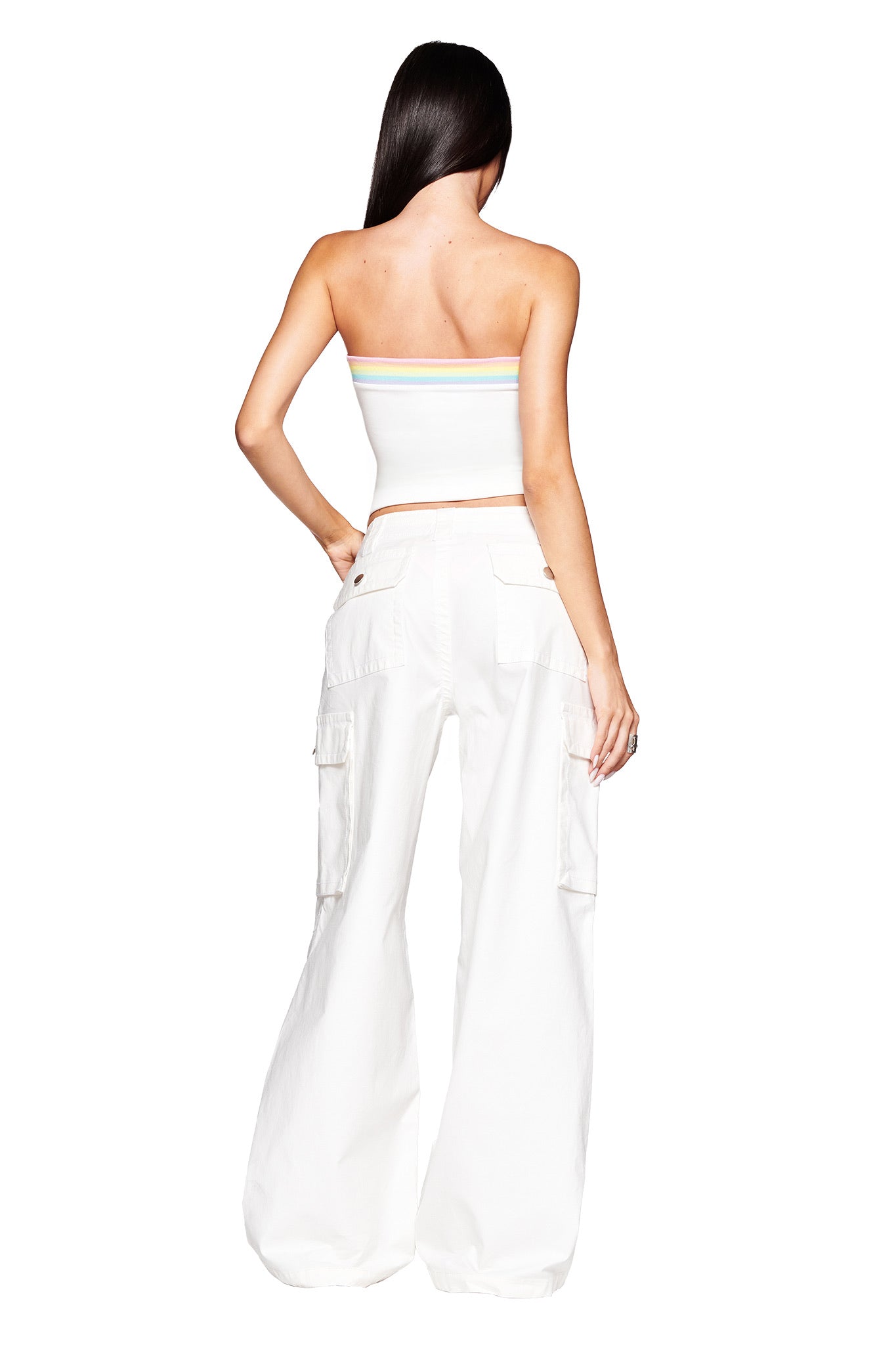 A woman with long, straight dark hair faces away from the camera, wearing the I.AM.GIA JAXXON PANT in white—featuring a baggy cargo silhouette—and a white strapless crop top with thin pastel stripes, set against a plain white background.