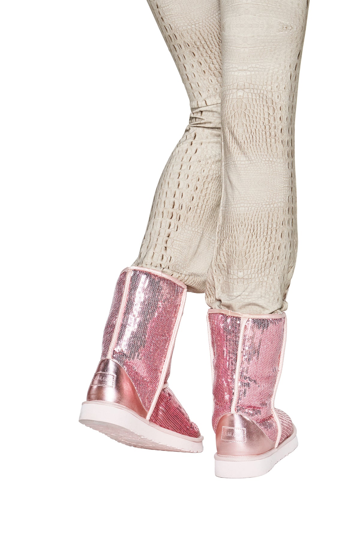 A person wearing textured beige pants and the I.AM.GIA ELVIRA SEQUIN BOOT in pink, photographed from behind against a white background.