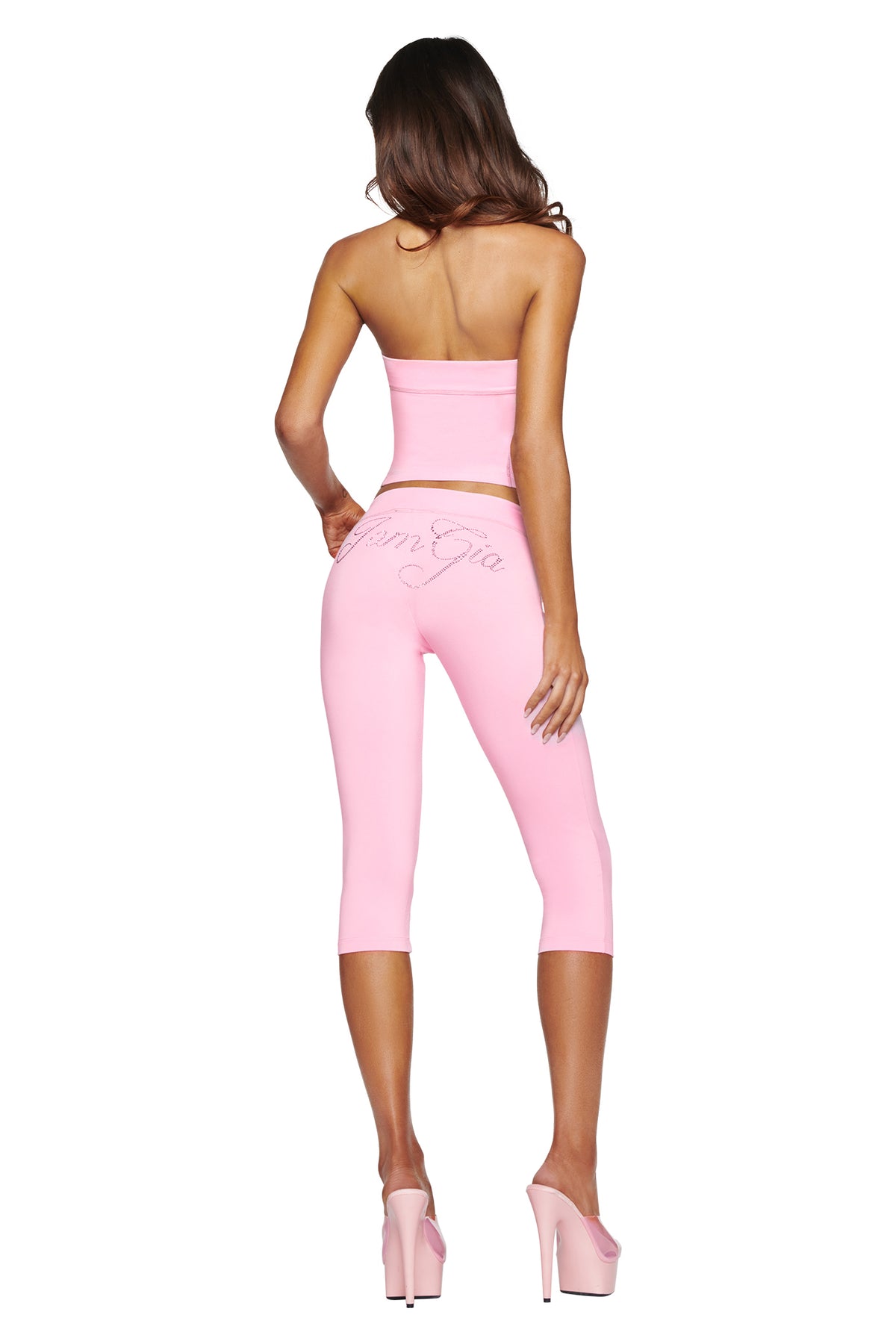 A woman stands facing away from the camera against a white background, wearing the I.AM.GIA BLARE DIAMONTE BOOB TUBE in baby pink, paired with pink capri leggings featuring a rhinestone logo script on the back and pink high heels.