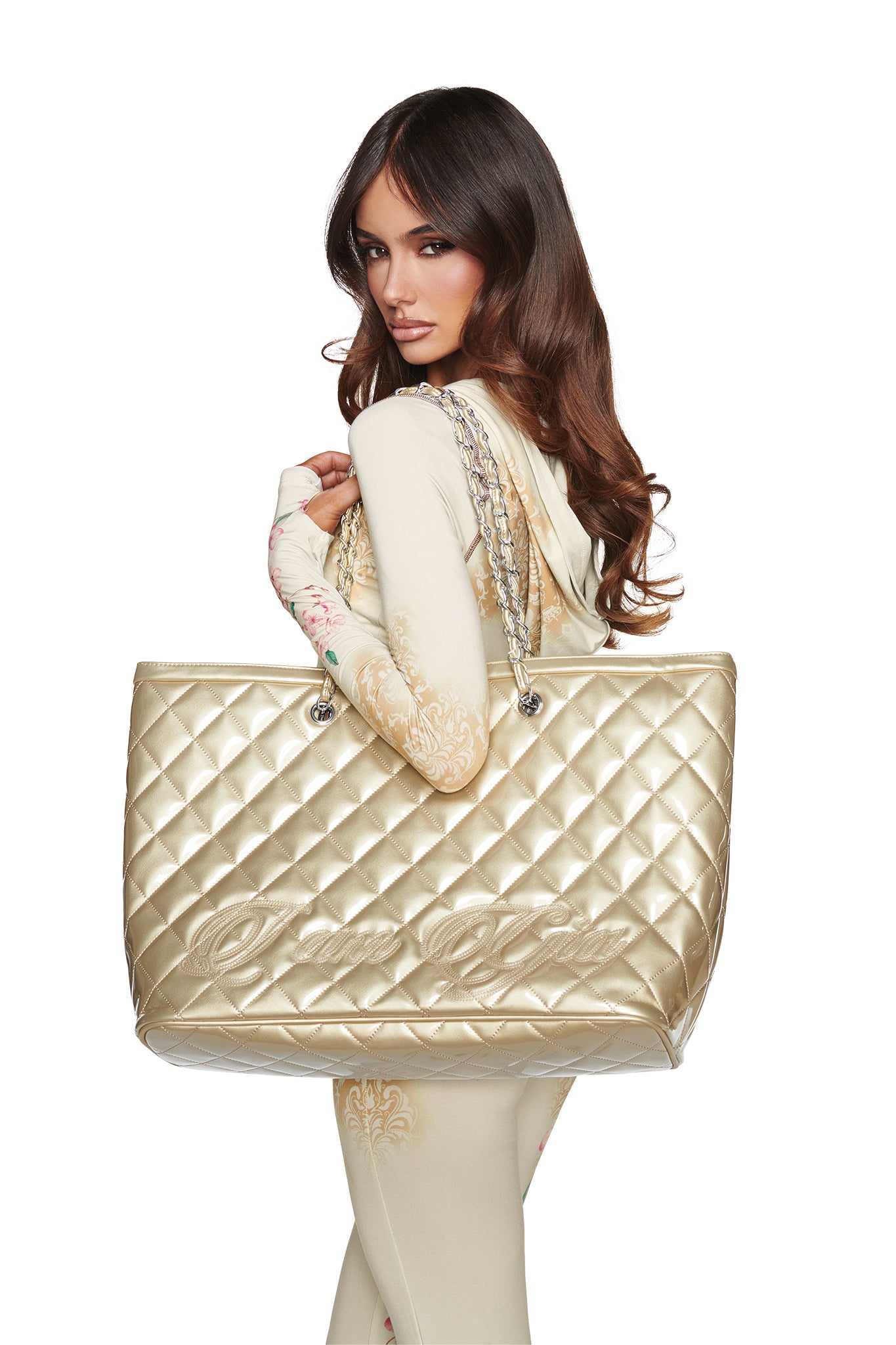 A woman dressed in a cream-colored outfit holds the I.AM.GIA DANNI TOTE BAG in gold, featuring oversized proportions and chain straps, as she looks over her shoulder against a white background.