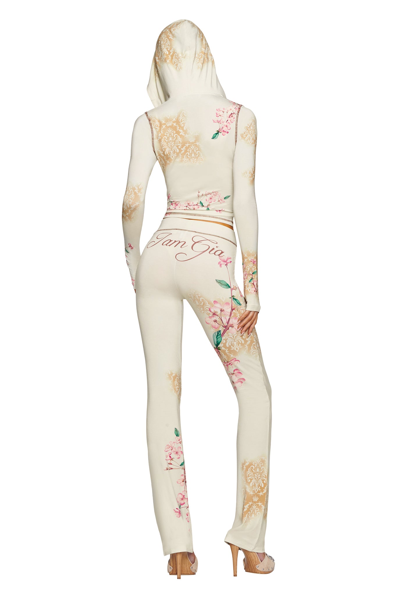 A person stands facing away, wearing the BLARE HOODIE - BLOSSOM and matching pants from I.AM.GIA, with “I.AM.GIA” printed on the back, paired with high heels.