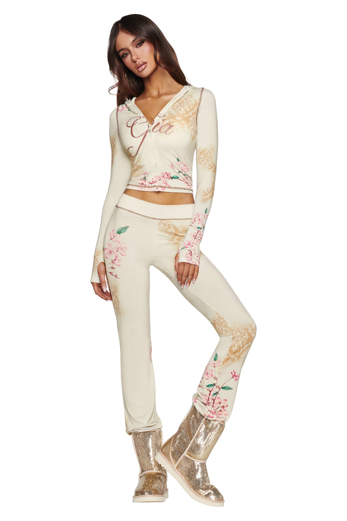 A woman poses in a cream floral two-piece outfit featuring a wrap top and matching pants. She completes the look with the I.AM.GIA ELVIRA SEQUIN BOOT in gold, standing stylishly against a white background.