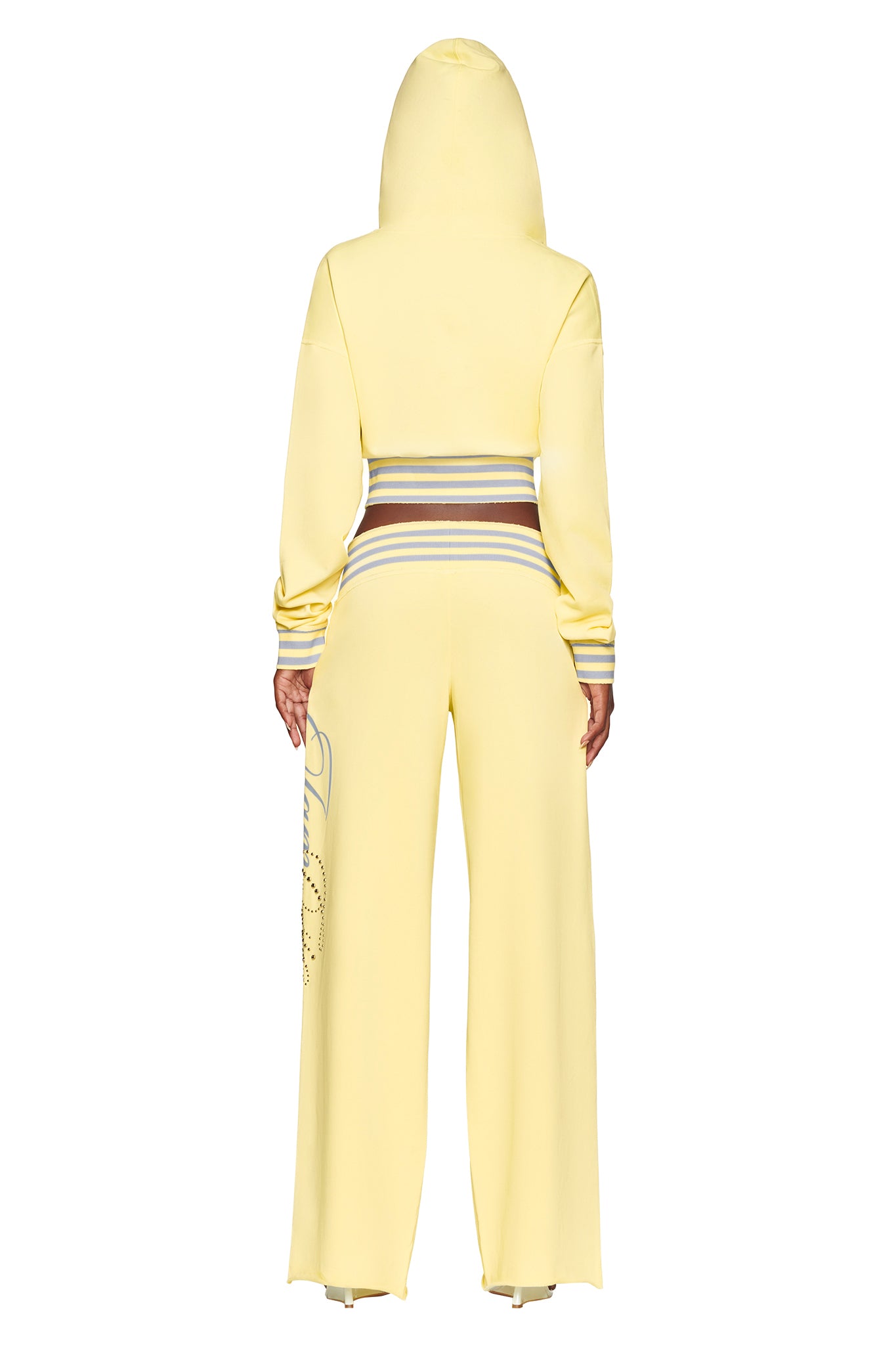A person is seen from the back wearing the I.AM.GIA VEDA PANT in yellow—a light yellow hooded tracksuit with a wide-leg silhouette. The outfit features multicolored stripes on the waistband and sleeves, and a graphic detail along one pant leg.