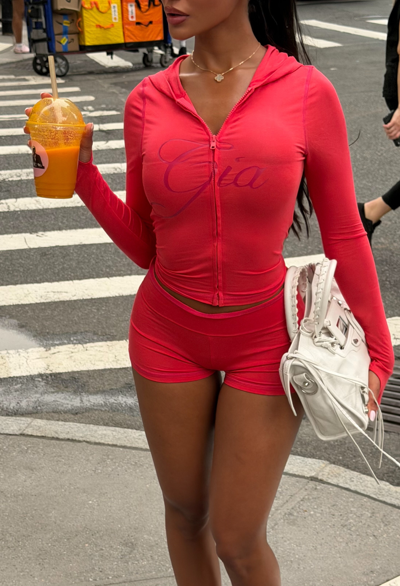 A woman wearing the I.AM.GIA BLARE HOODIE in Papaya and matching shorts stands on a city street, holding an orange drink and a white handbag. Only her body is visible; her face is not shown.