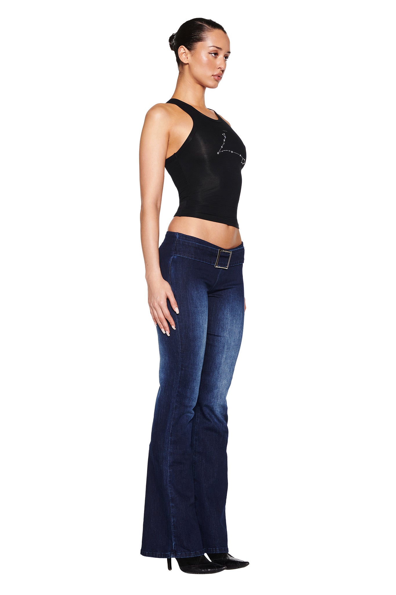 A person stands sideways against a white backdrop, wearing an I.AM.GIA black sleeveless top and SHARAH DENIM PANT - BLUE, dark blue flared jeans with a belt. Their hair is pulled back, complemented by black heels. The jeans have a low-rise waist, adding modern flair to the classic look.