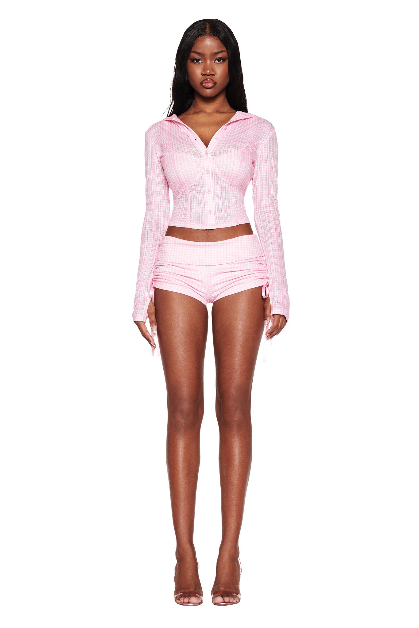 A woman stands facing forward, wearing the I.AM.GIA MIA SHORT - PINK GINGHAM long-sleeve top and high-waisted shorts set. Her hair is long and dark, complemented by clear high heels against a pristine white background.