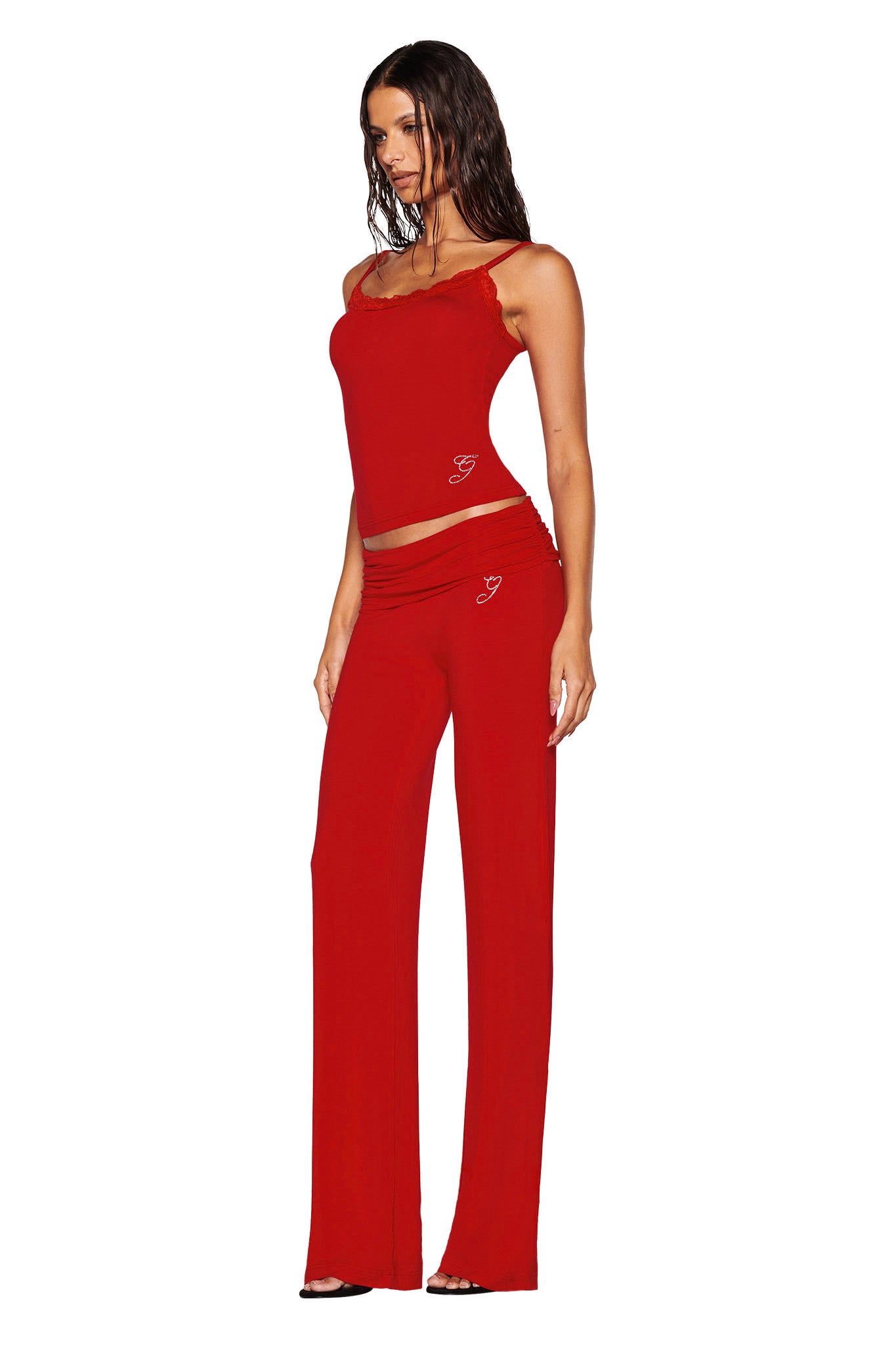 A woman wearing the I.AM.GIA ERIN TOP in red, paired with matching pants, poses against a white background. Her long dark hair elegantly cascades as she complements the outfit with heels. The ensemble, crafted from bamboo fabric, is adorned with a subtle heart design that enhances its charm.