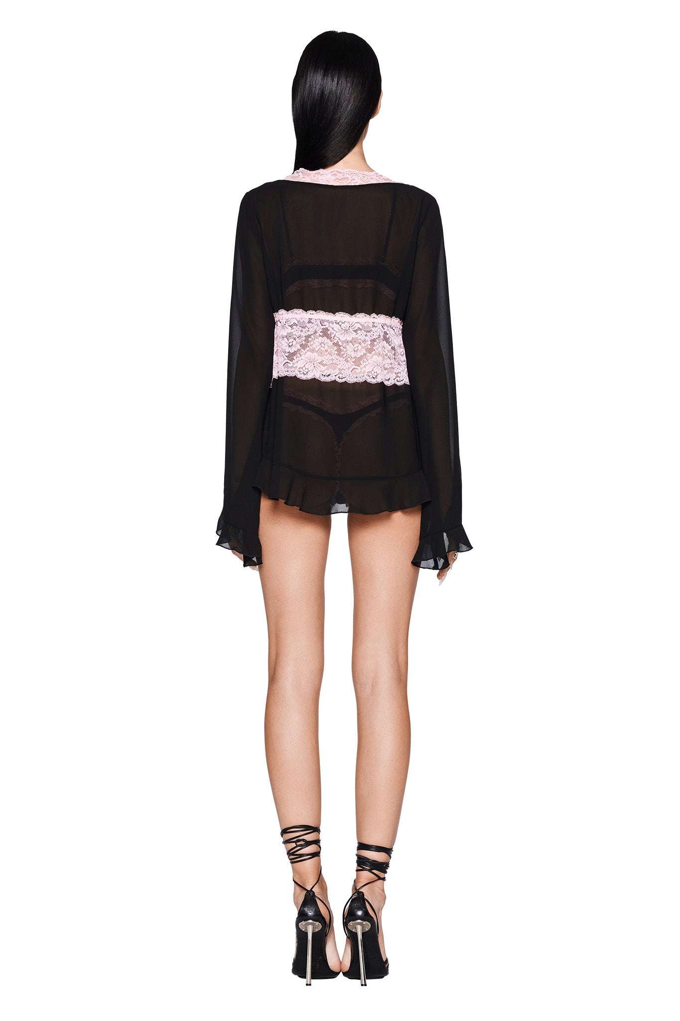A person with long black hair stands facing away from the camera, wearing the JUNIPER ROBE THREE-PIECE SET - BLACK by I.AM.GIA. A wide band of contrasting pink lace encircles their waist, perfectly matching their strappy high-heeled sandals. The set's pull-on construction offers a chic and effortless look against a plain white backdrop.