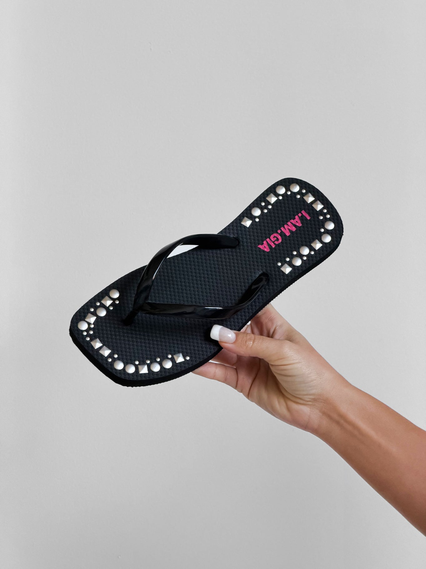 A hand with light skin holds the I.AM.GIA INKA STUDDED FLIP FLOP in black, detailed with white pearls along the edge and "JAMAICA" in pink on the sole, against a plain light gray background.