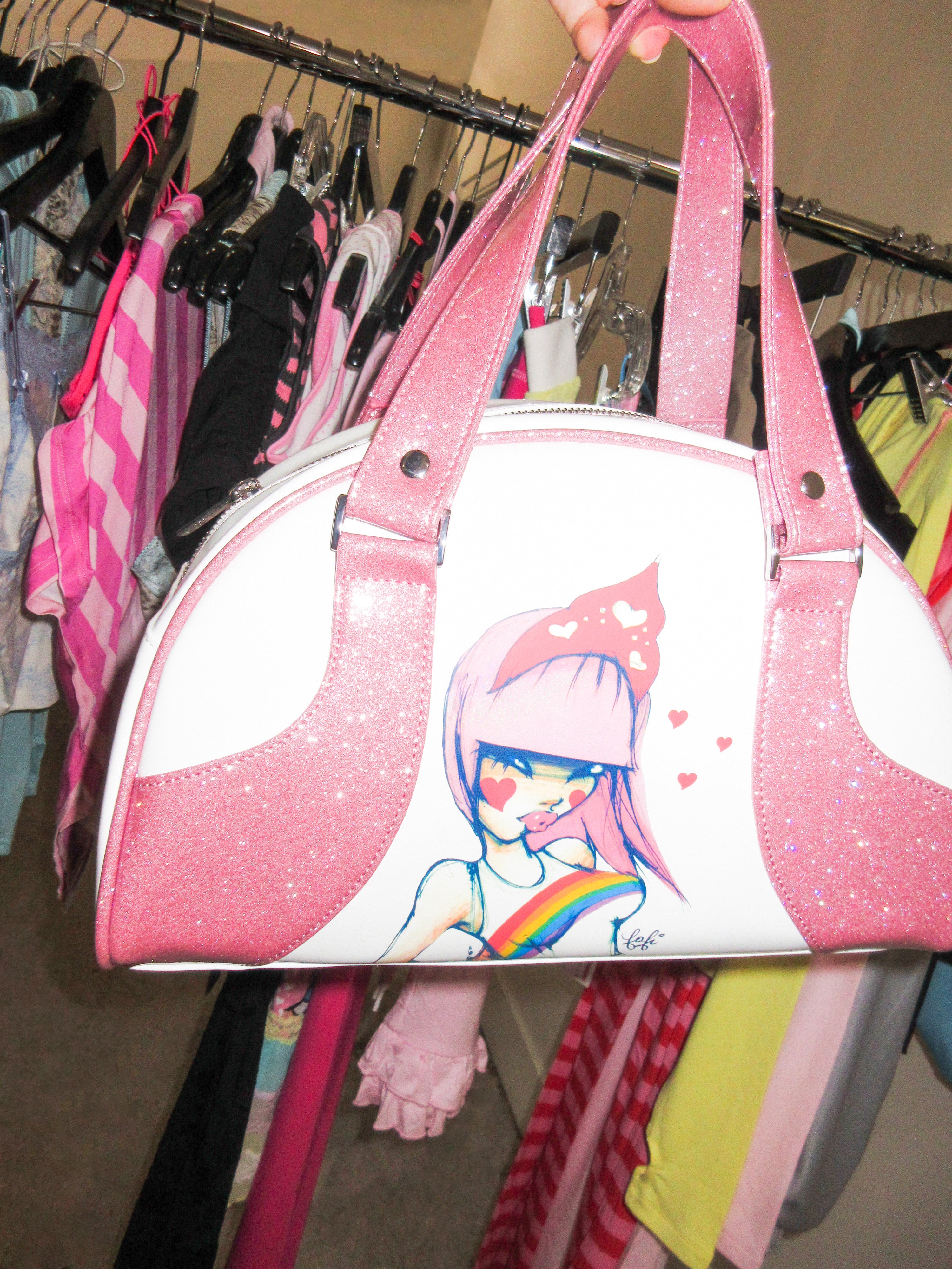 The FAFI BAG - PINK by I.AM.GIA, featuring glittery handles and a colorful girl illustration, is held before a vibrant clothing rack—a playful accessory for any FAFI collaboration fan.