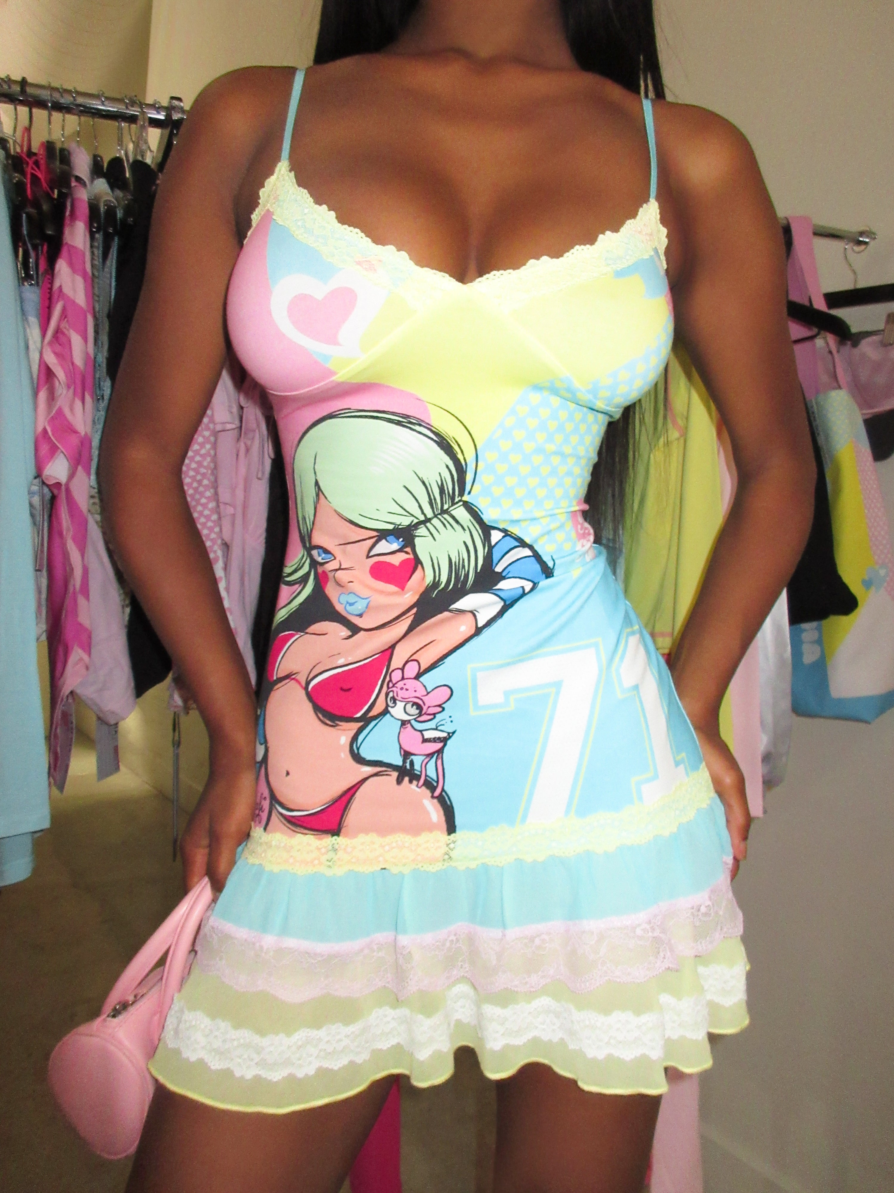 A woman stands in a closet wearing the I.AM.GIA FAFI MINI DRESS - BLUE, a stretch jersey cami mini dress with an exclusive graphic print featuring a cartoon woman and the number 71. Clothes and a pink purse are visible in the background.