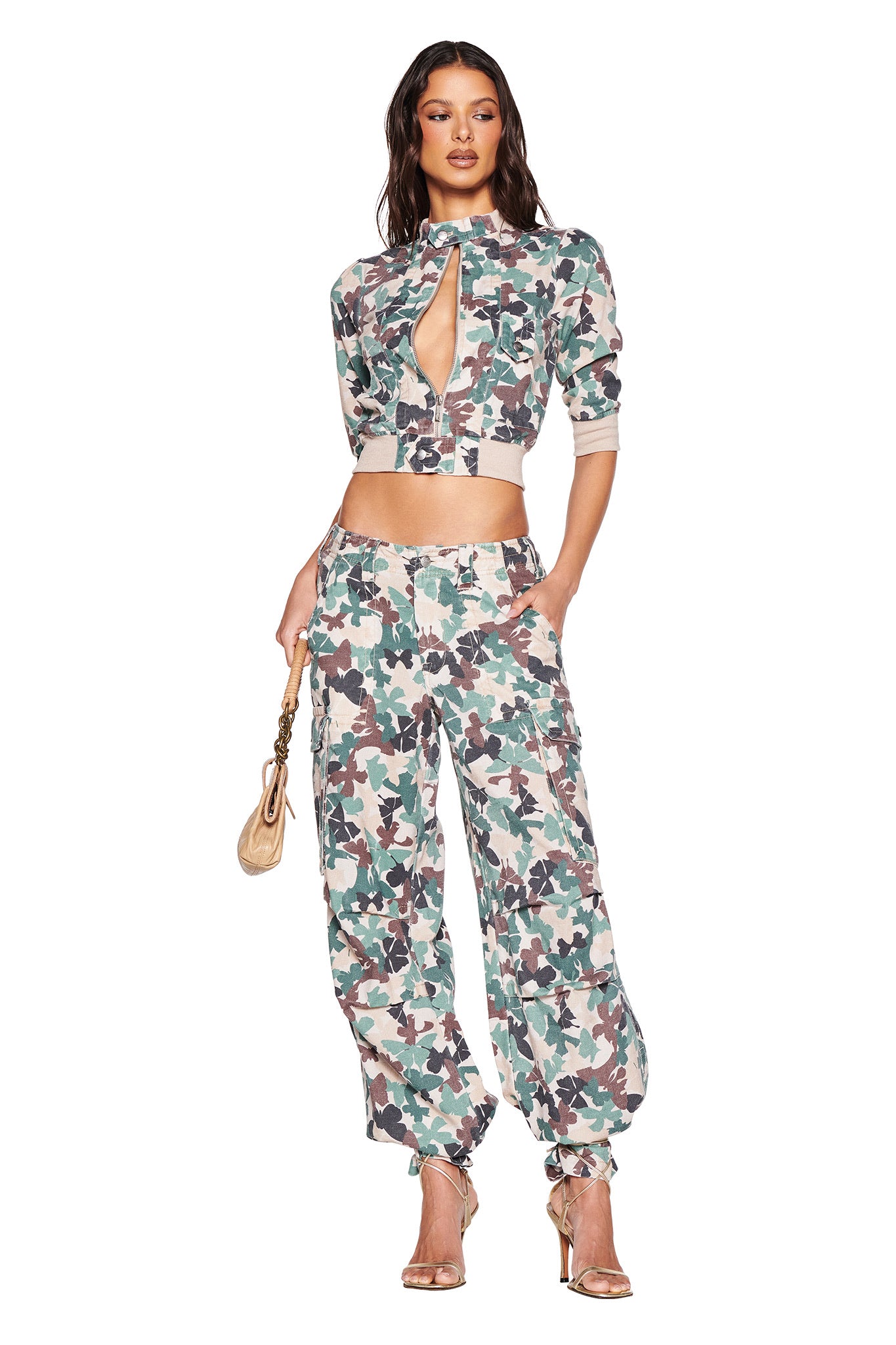 A person poses confidently in I.AM.GIA's EFFIE CARGO PANT - FLUTTER CAMO with a matching top featuring a keyhole cutout while holding a small tan purse. Gold high heels add elegance to the look.