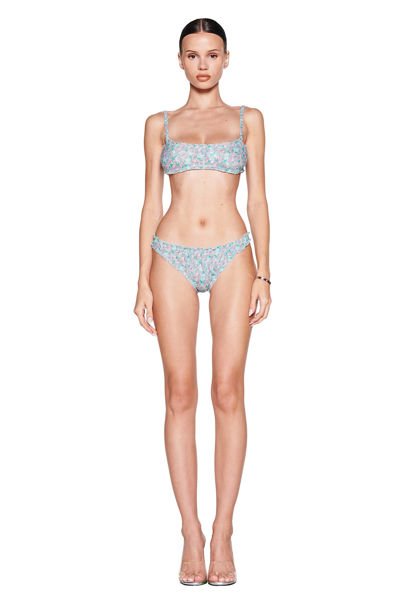 A person stands facing forward in a vintage-inspired I.AM.GIA "JEAN BRALETTE SET - TEAL BUTTERFLY BLOOM," featuring a graceful butterfly print on light blue floral fabric. Their straight hair is pulled back, and they complete the look with clear high-heeled shoes.