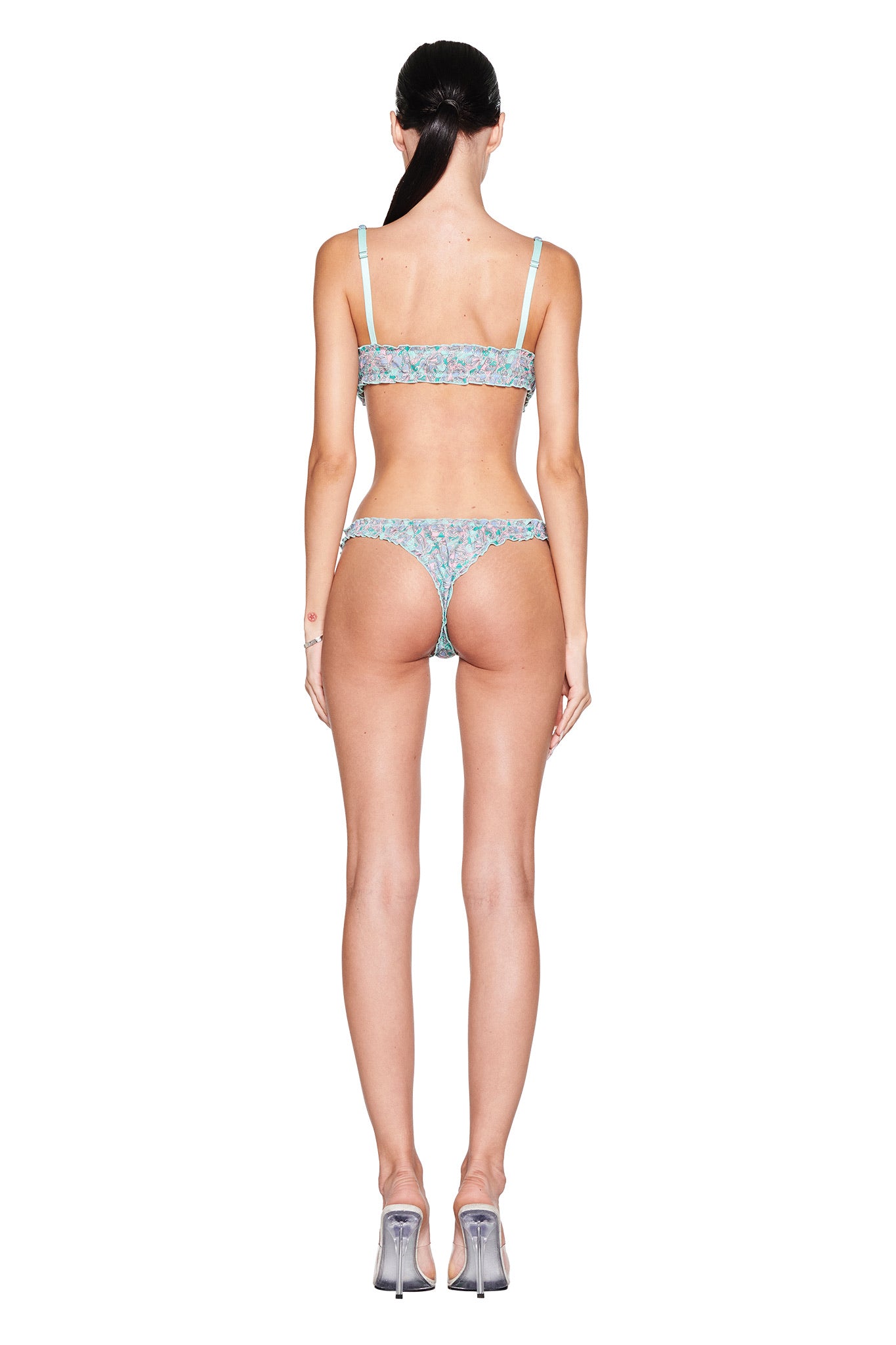 Facing away from the camera on a white backdrop, they wear I.AM.GIA's JEAN BRALETTE SET in TEAL BUTTERFLY BLOOM and transparent heels. Their long dark hair is tied back, enhancing the vintage-inspired butterfly motif's whimsical appeal.