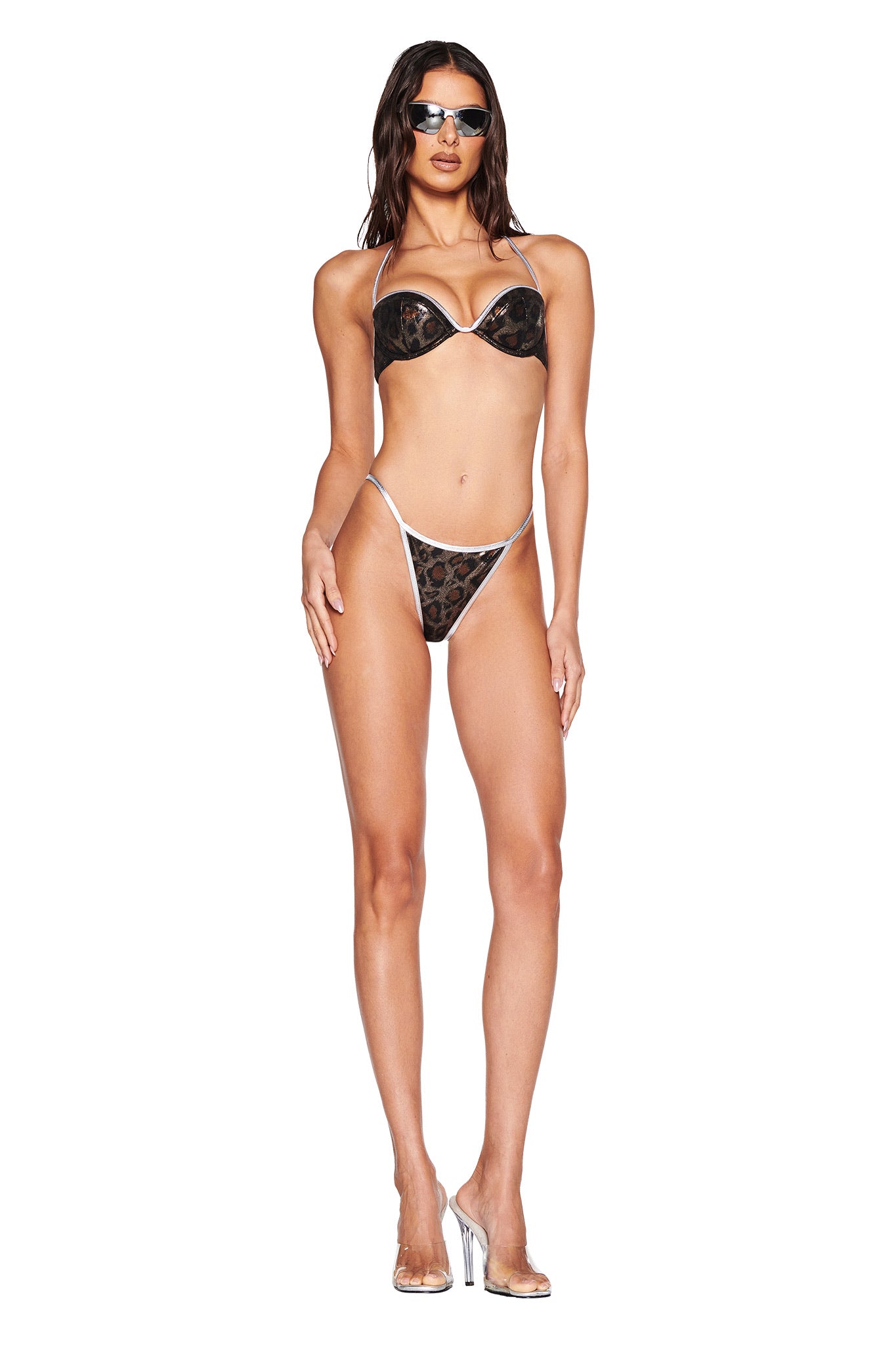 A woman poses against a white background in the I.AM.GIA HONEY BIKINI TOP - CLASSIC LEOPARD. She has long dark hair, sunglasses, and clear high heels, accentuating the bold lurex fabric.