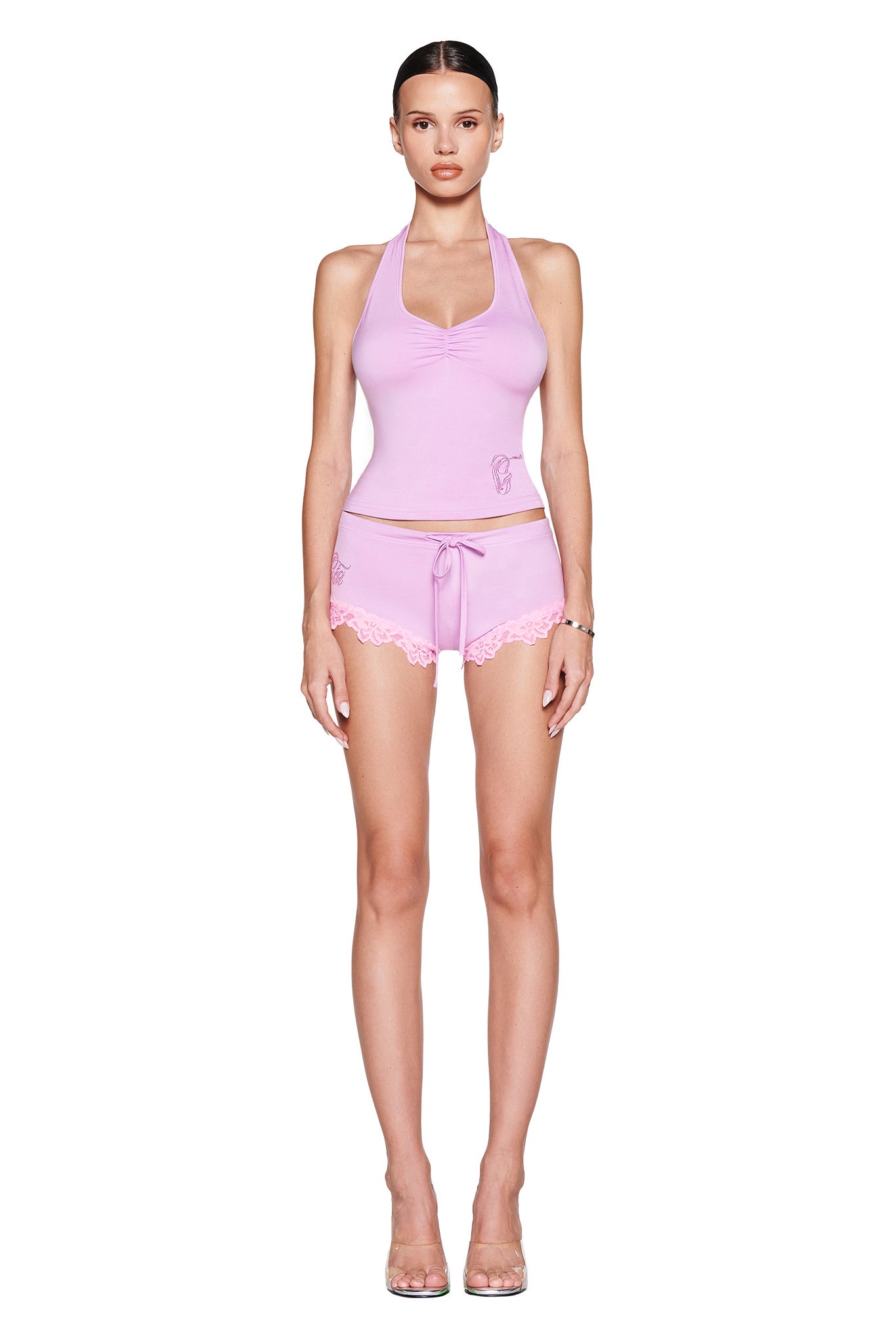 A person, with long hair tied back, wears a stylish light pink halter top and matching lace-trimmed micro mini skirt. Clear high heels complete the look, showcased against a lavender background complementing the CELESTE SHORT - LAVENDER from I.AM.GIA.