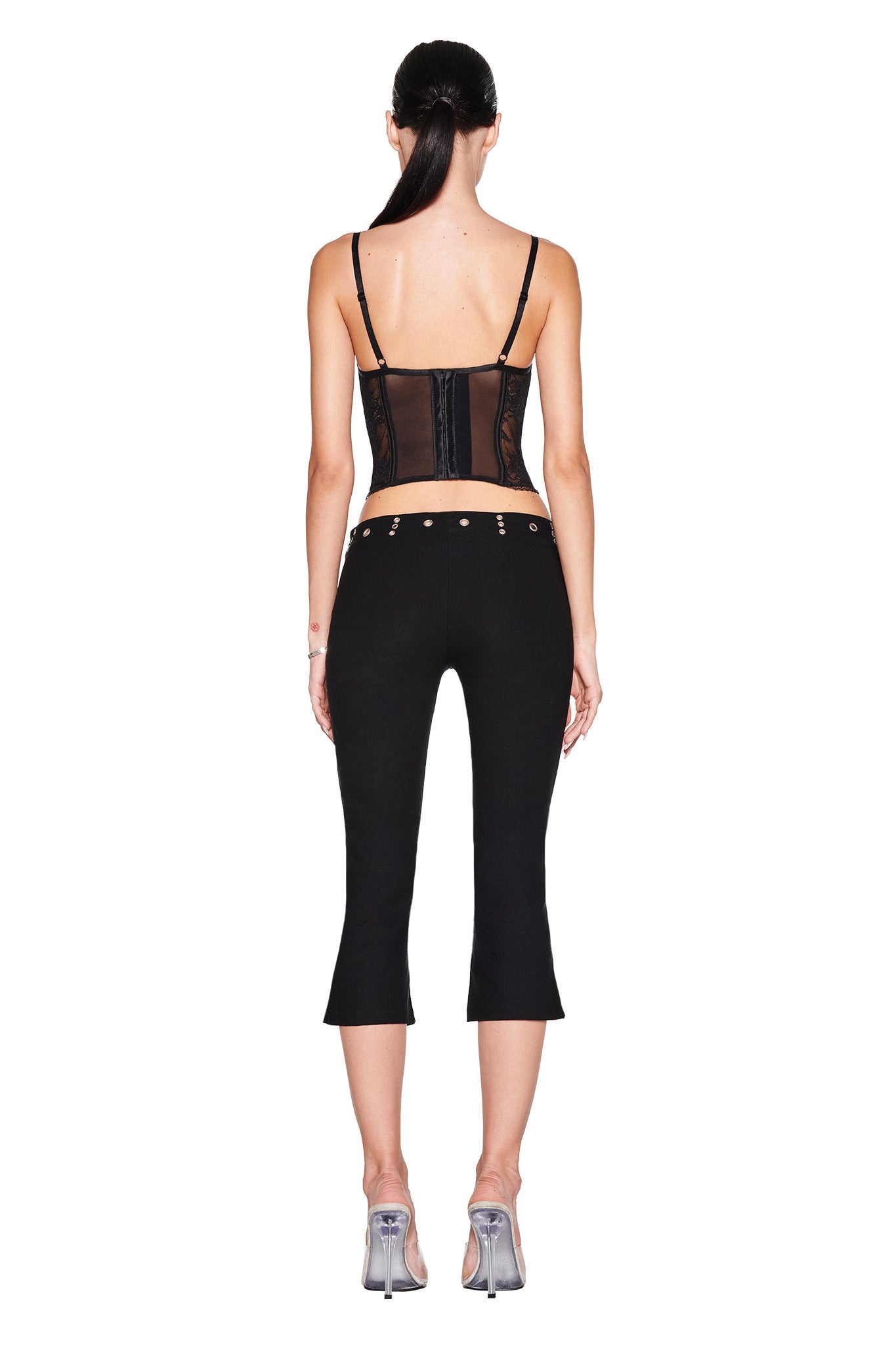 A woman faces away, wearing the I.AM.GIA SHARAH CAPRI - BLACK low-rise, flared pants with a slim black corset top and clear heels. Her dark hair is in a low ponytail against a white background.