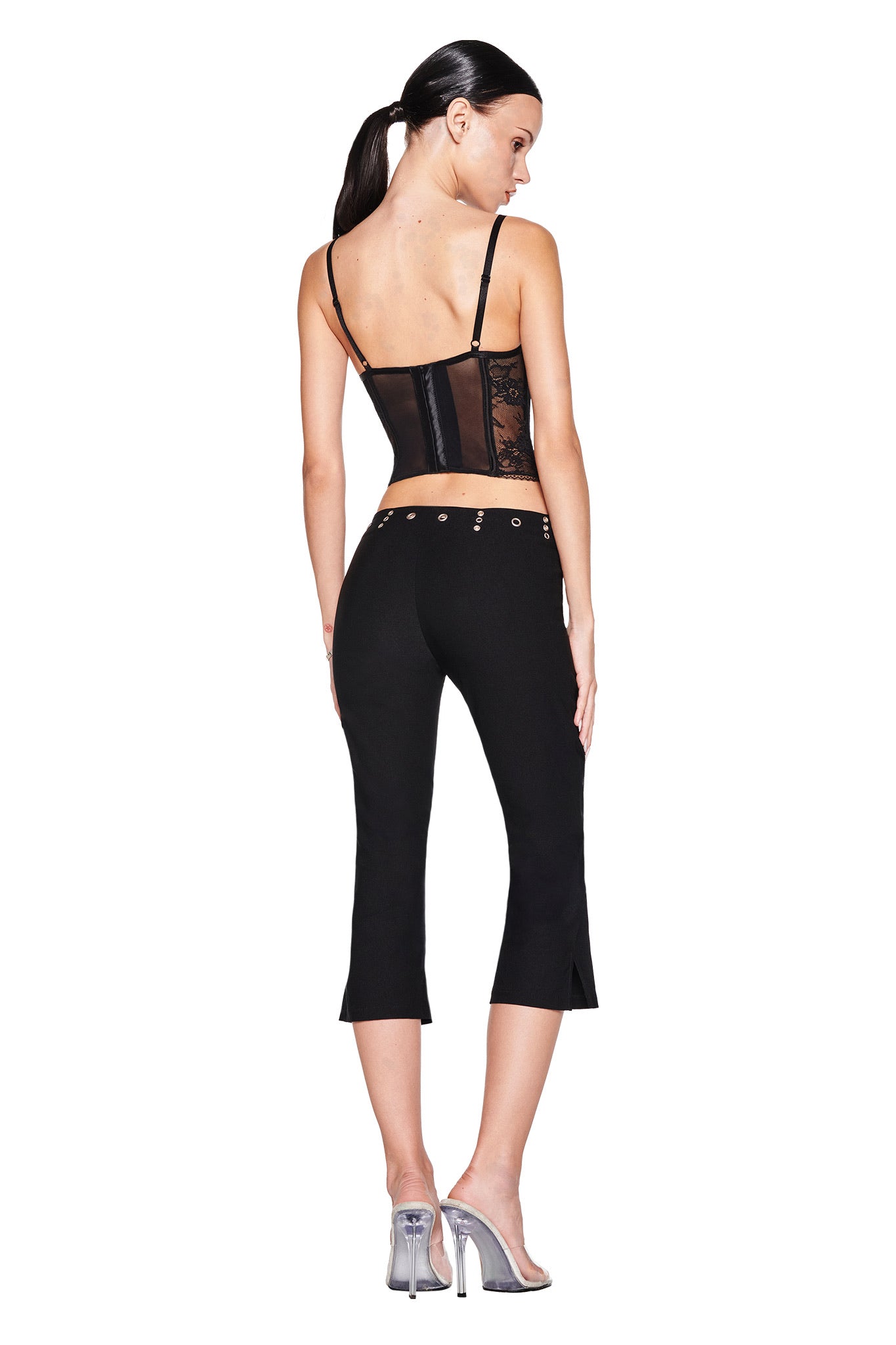 A woman with a ponytail, facing away, models the I.AM.GIA SHARAH CAPRI - BLACK: slim-fit, low-rise black capri pants with grommet details, paired with a sheer black corset top and clear heels against a white backdrop.