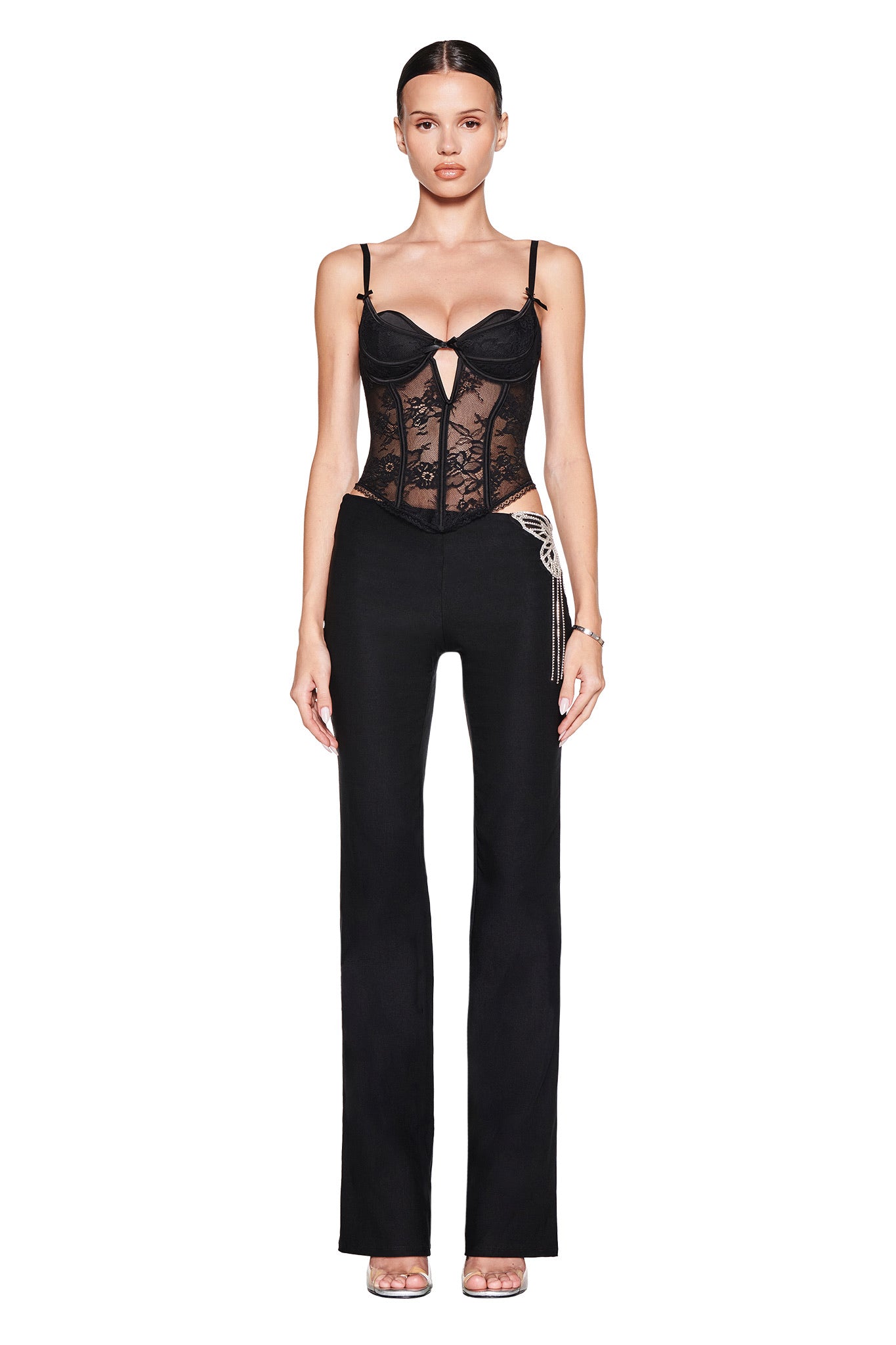 A woman faces forward against a white background, wearing I.AM.GIA's SORREL PANT in black and a black lace corset top with spaghetti straps. Her hair is slicked back and she pairs the look with open-toed heels.