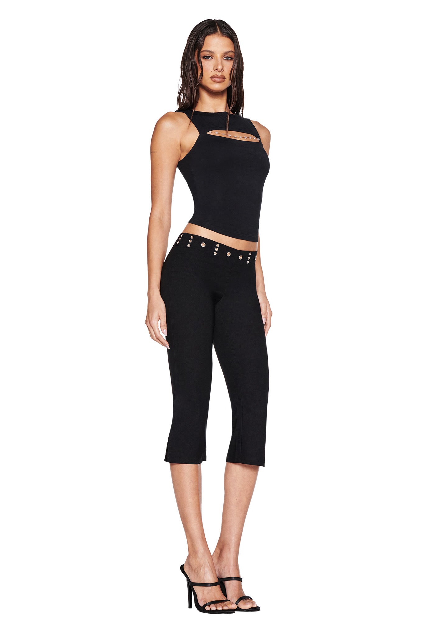 A woman poses against a white background wearing the I.AM.GIA SHARAH CAPRI - BLACK, featuring slim fit and silver-studded low-rise waistband, styled with a sleeveless black halter top and black high-heeled sandals.
