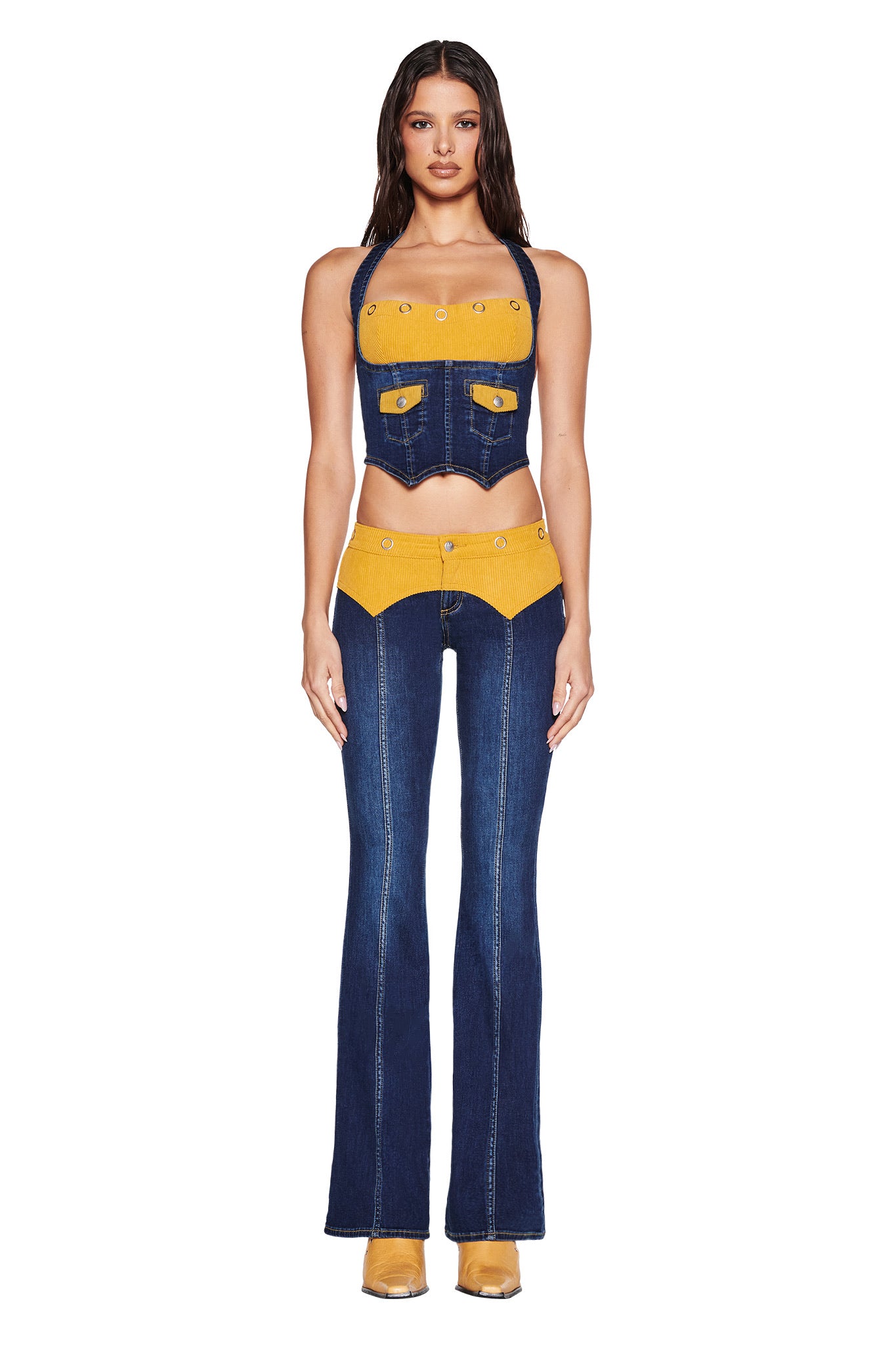 A woman faces forward in I.AM.GIA’s TOMMIE JEAN - DARK WASH flared denim pants with a mustard corduroy waistband and a sleeveless crop top with GIA logo embroidery. She has long loose hair, tan shoes, and stands against a white background.