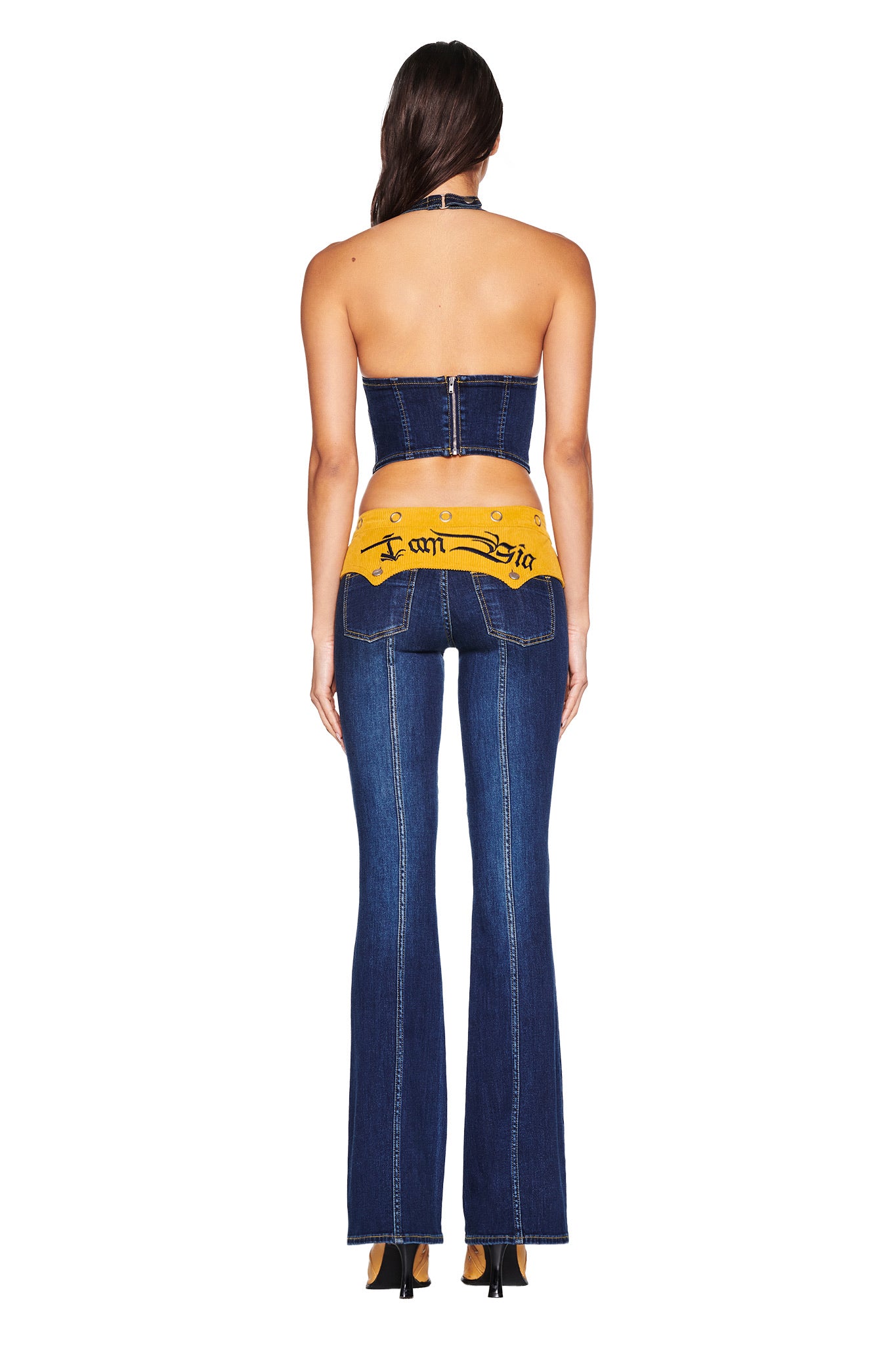 A woman faces away, wearing I.AM.GIA’s TOMMIE JEAN - DARK WASH: a dark denim halter crop top and flared pants with a mustard yellow corduroy waistband and black script, paired with high heels.