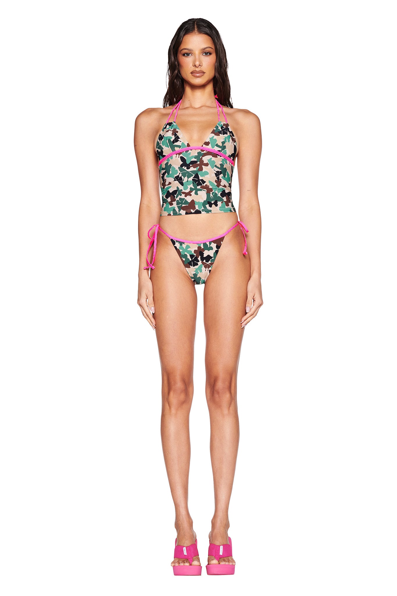 The person is wearing the I.AM.GIA SEDONA TANKINI in Flutter Camo with neon pink bindings, matching side-tie bottoms, and pink platform sandals, creating a standout look against the plain white background.