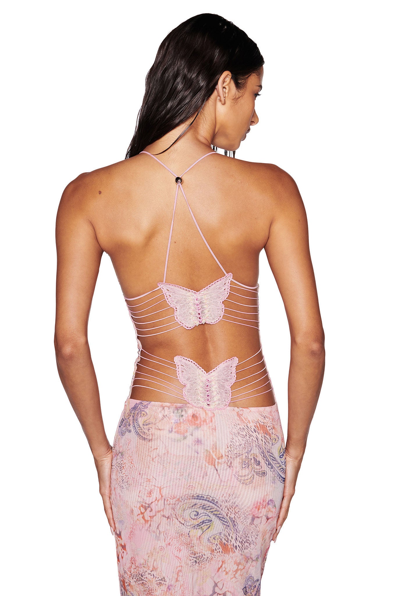 A person with long, dark hair stands with their back facing the camera, wearing the ELLERY DRESS - PINK : FLORA PAISLEY from I.AM.GIA. The halter neck style dress features intricate butterfly designs and thin straps crisscrossing their exposed open back. The outfit is paired with a patterned, flowy skirt.