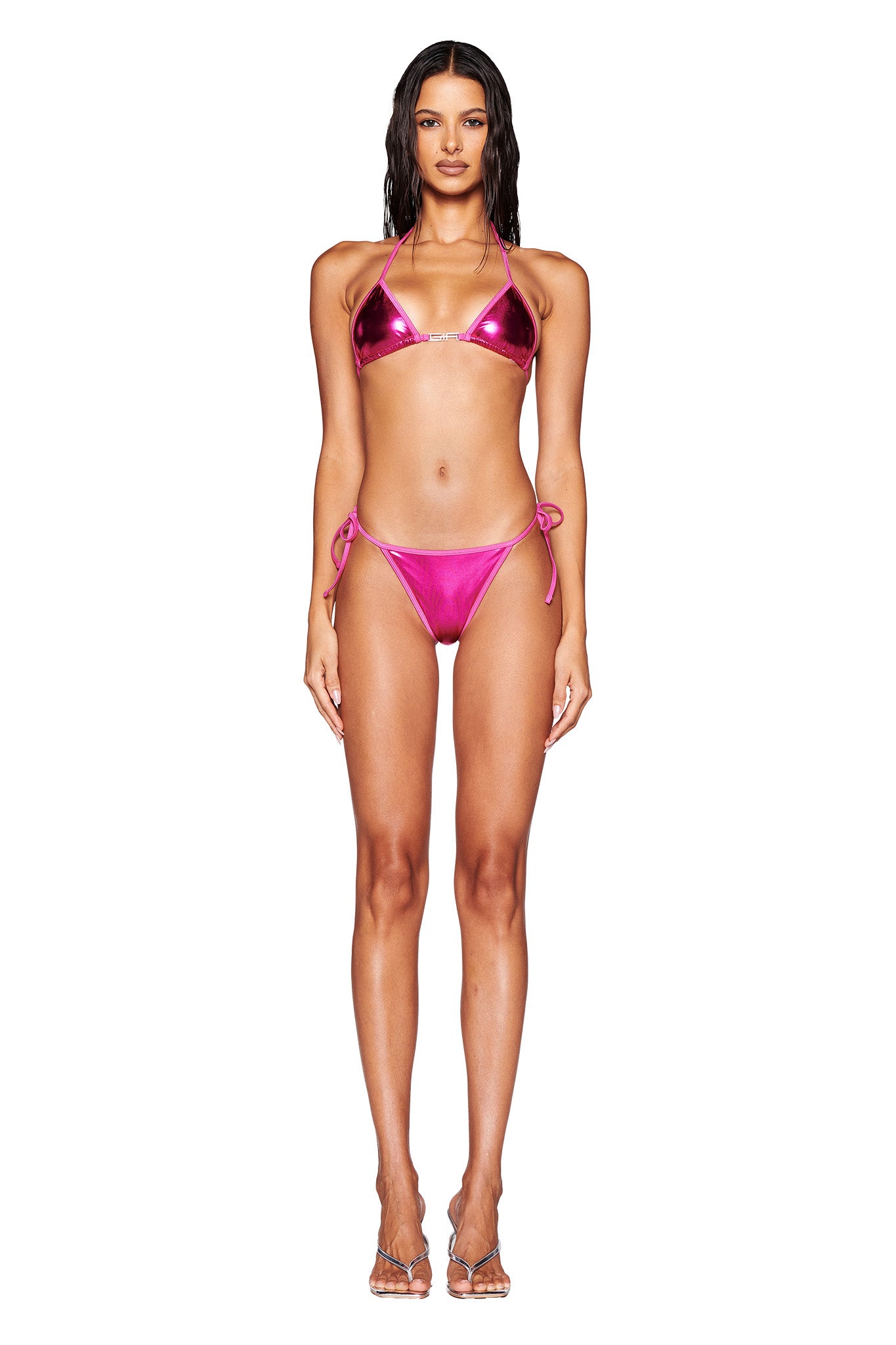 A woman with long hair stands facing forward, wearing the I.AM.GIA Alohi Bikini Bottom in hot pink, which features a shiny, metallic finish. She sports a halter neck top and cheeky bikini bottom with tie-sides. She completes her look with clear high-heeled shoes. The background is white and free of any additional elements.