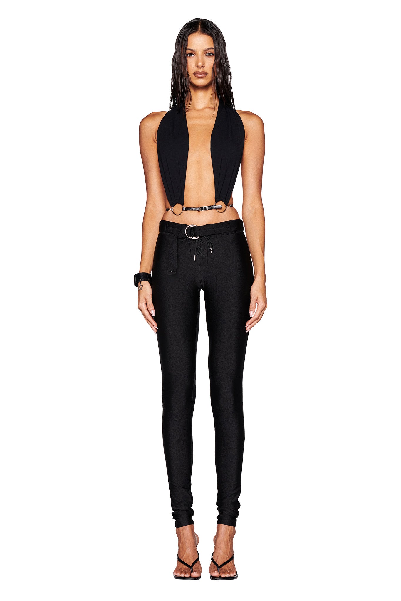A person stands against a white background wearing the MIKAYLA TOP - BLACK by I.AM.GIA, featuring a deep neckline and paired with black fitted pants. A faux leather belt cinches the look perfectly. They have long, wet-look hair and are wearing strappy black heels.