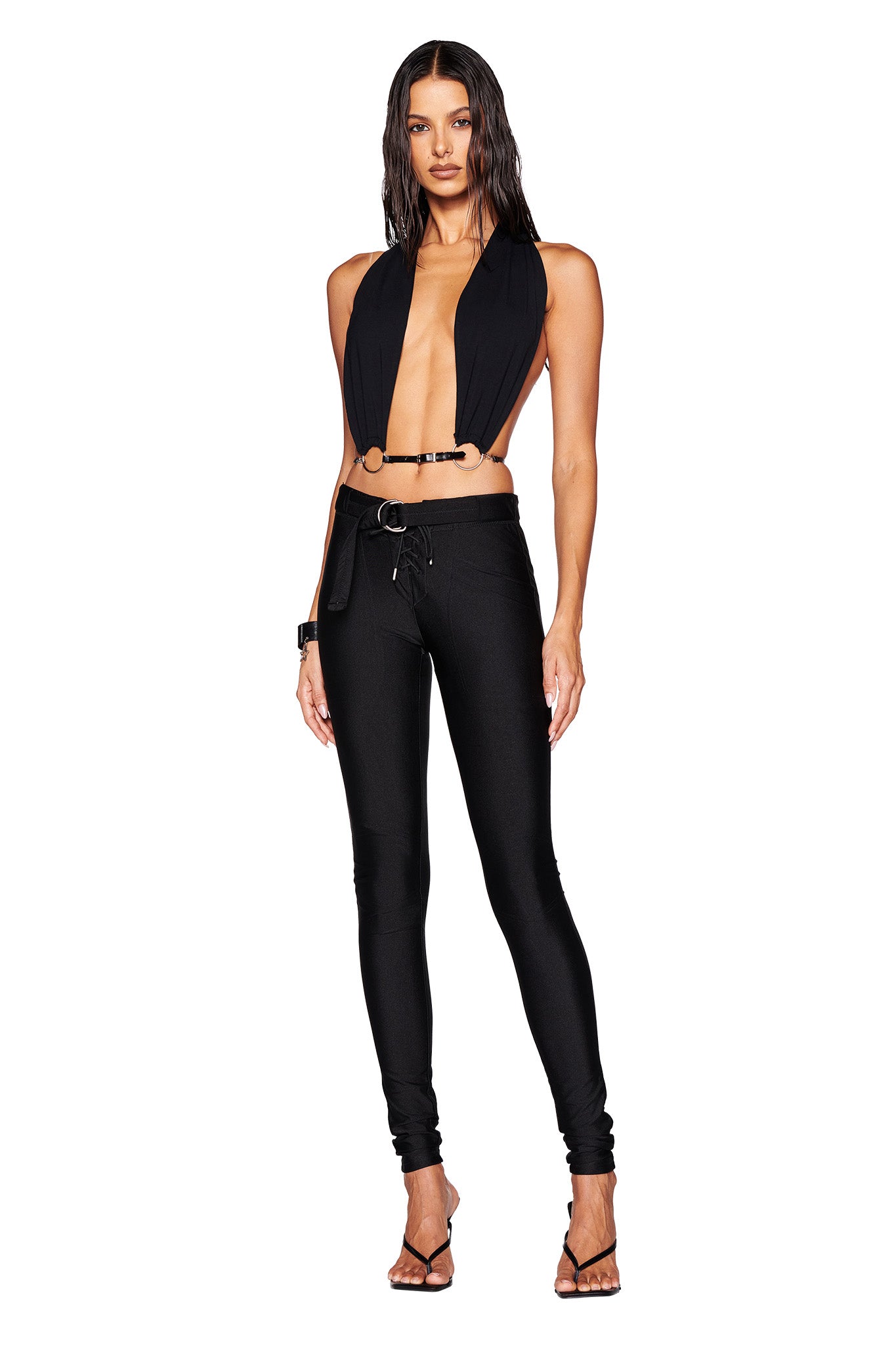 A woman with long dark hair confidently wears the I.AM.GIA MIKAYLA TOP in black, paired with high-waisted pants cinched by a faux leather belt. Her strappy high heels perfectly complement the sleek ensemble against a plain white background.