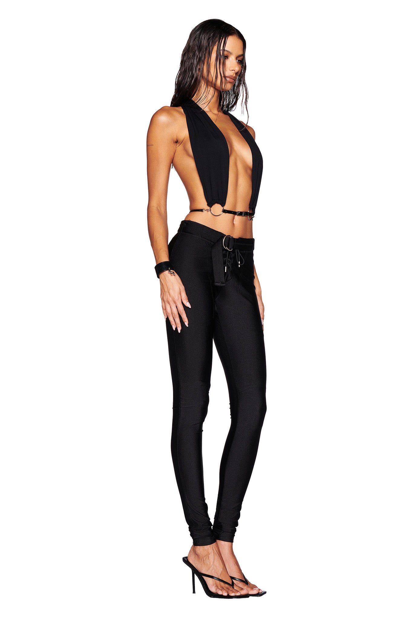 A person stands in a side profile pose, wearing the MIKAYLA TOP in black by I.AM.GIA paired with sleek black pants. They have long, wet-looking hair, black high-heeled sandals, and a confident expression accentuated by a chic faux leather belt.