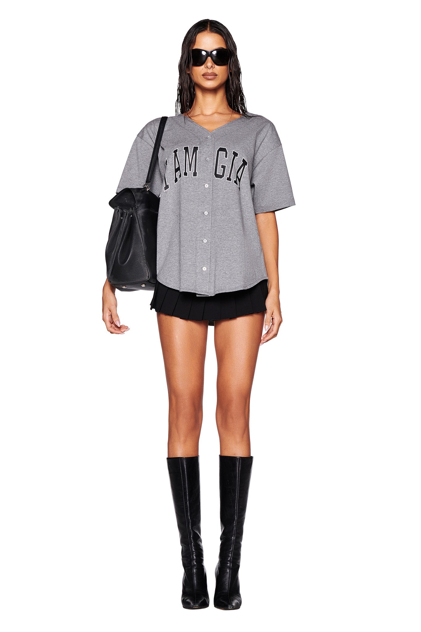 A person stands against a white background wearing black sunglasses, the Lorri Jersey in steel grey and black from I.AM.GIA, a pleated black mini skirt, black knee-high boots, and a large black handbag on their shoulder.