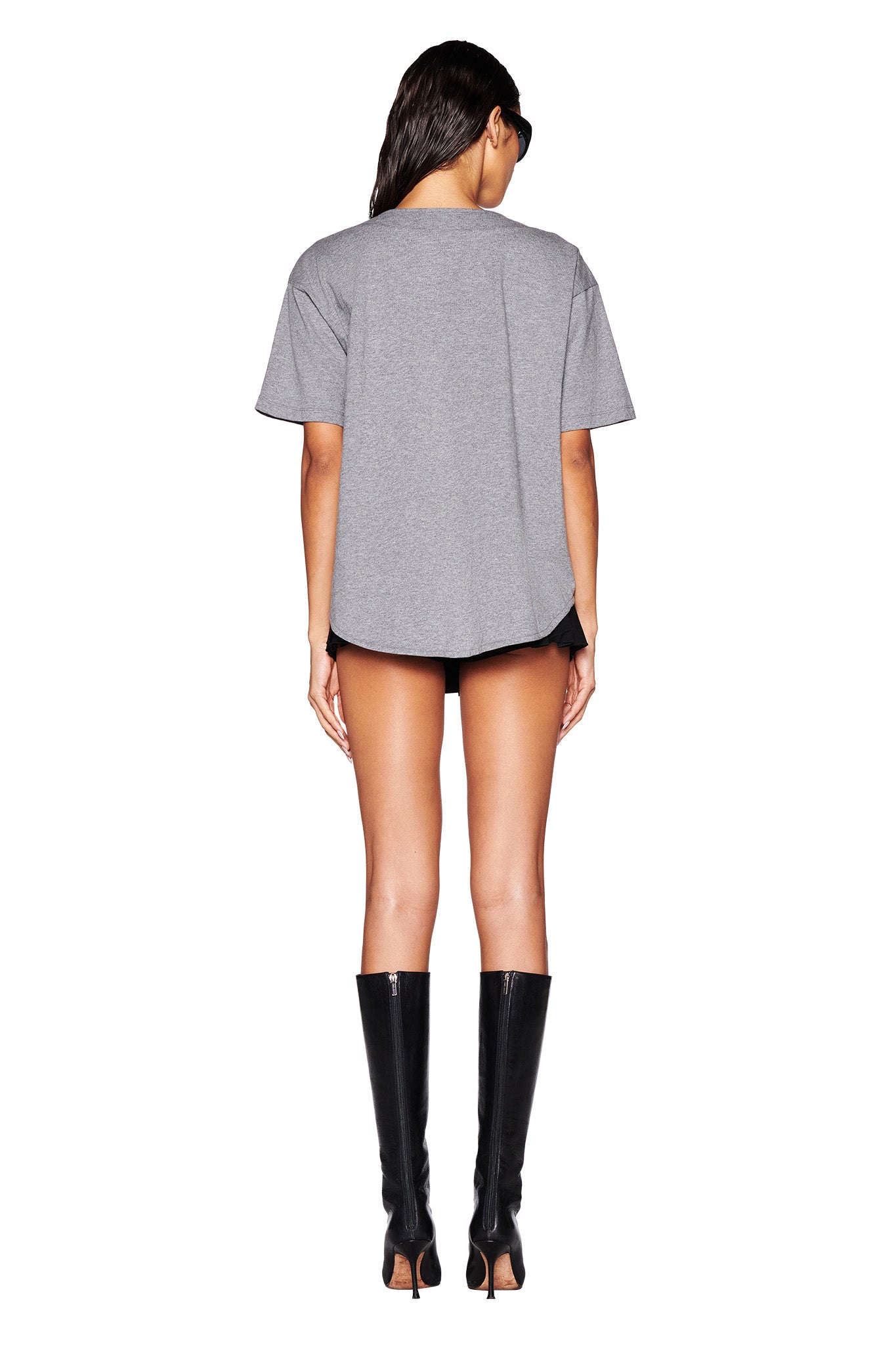A person with long hair is standing with their back to the camera in an oversized LORRI JERSEY in steel grey from I.AM.GIA, paired with black shorts and high-heeled black knee-high boots. They're also wearing sunglasses against a plain white background.