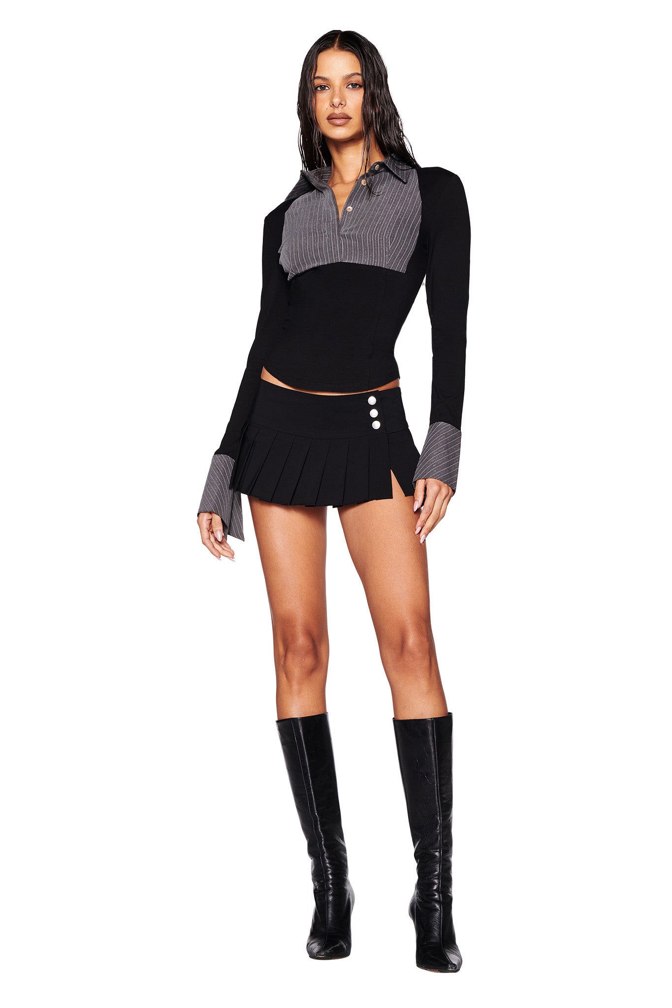 A woman stands confidently wearing the I.AM.GIA SHARAH TOP in black and grey, layered over a pinstripe button-up shirt, paired with a black pleated mini skirt featuring button details. She completes her look with black knee-high boots, and her hair is styled in loose waves.