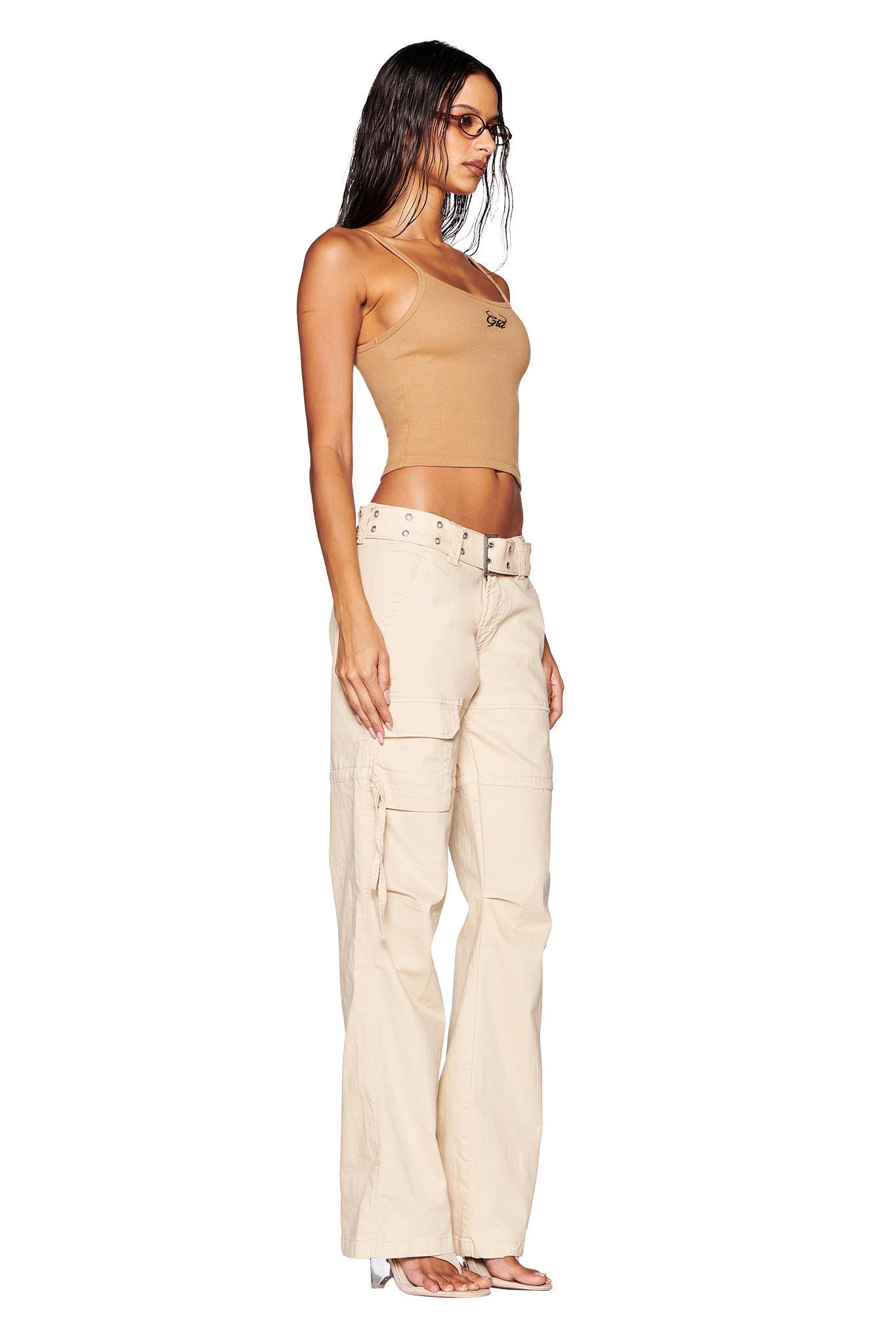 A person stands in a side profile pose, wearing a beige tank top, 100% cotton NAIMA CARGO PANT - TAN by I.AM.GIA with a white belt and clear-heeled sandals. They have long, dark hair and sunglasses against a plain white background.