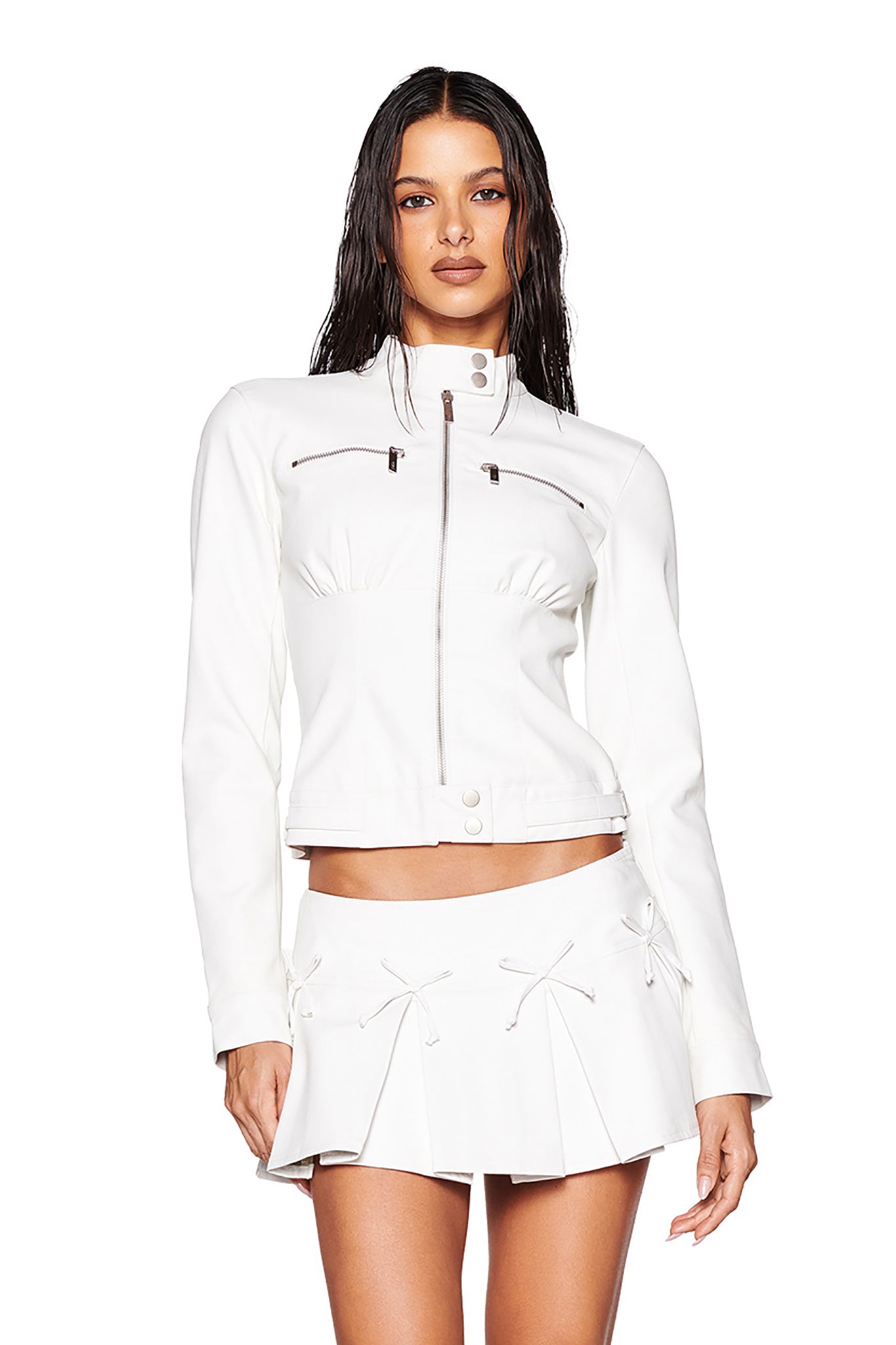 A woman with long, wet hair is wearing the stylish EVERETTE JACKET in white by I.AM.GIA, featuring a zip-up front and two small chest pockets. The slimming fit of the jacket beautifully complements her matching pleated white skirt. This chic, modern ensemble stands out against a plain background.