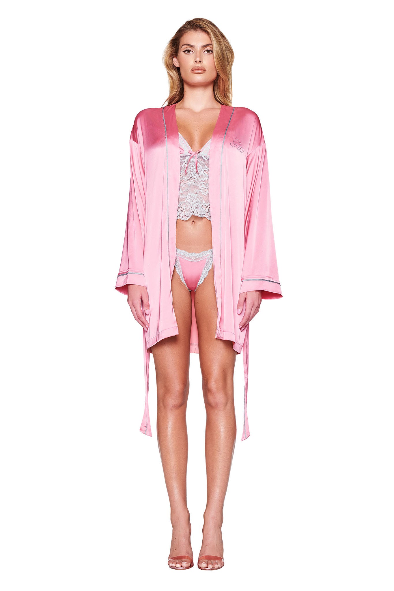 A person stands against a plain white background, wearing the MARTINA SLEEP ROBE in pink by I.AM.GIA. The robe, featuring custom 'GIA' diamante detailing, is open to reveal a matching lace lingerie set and high heels beneath. The robe's silky satin material drapes elegantly in a relaxed fit as the individual showcases their long hair.