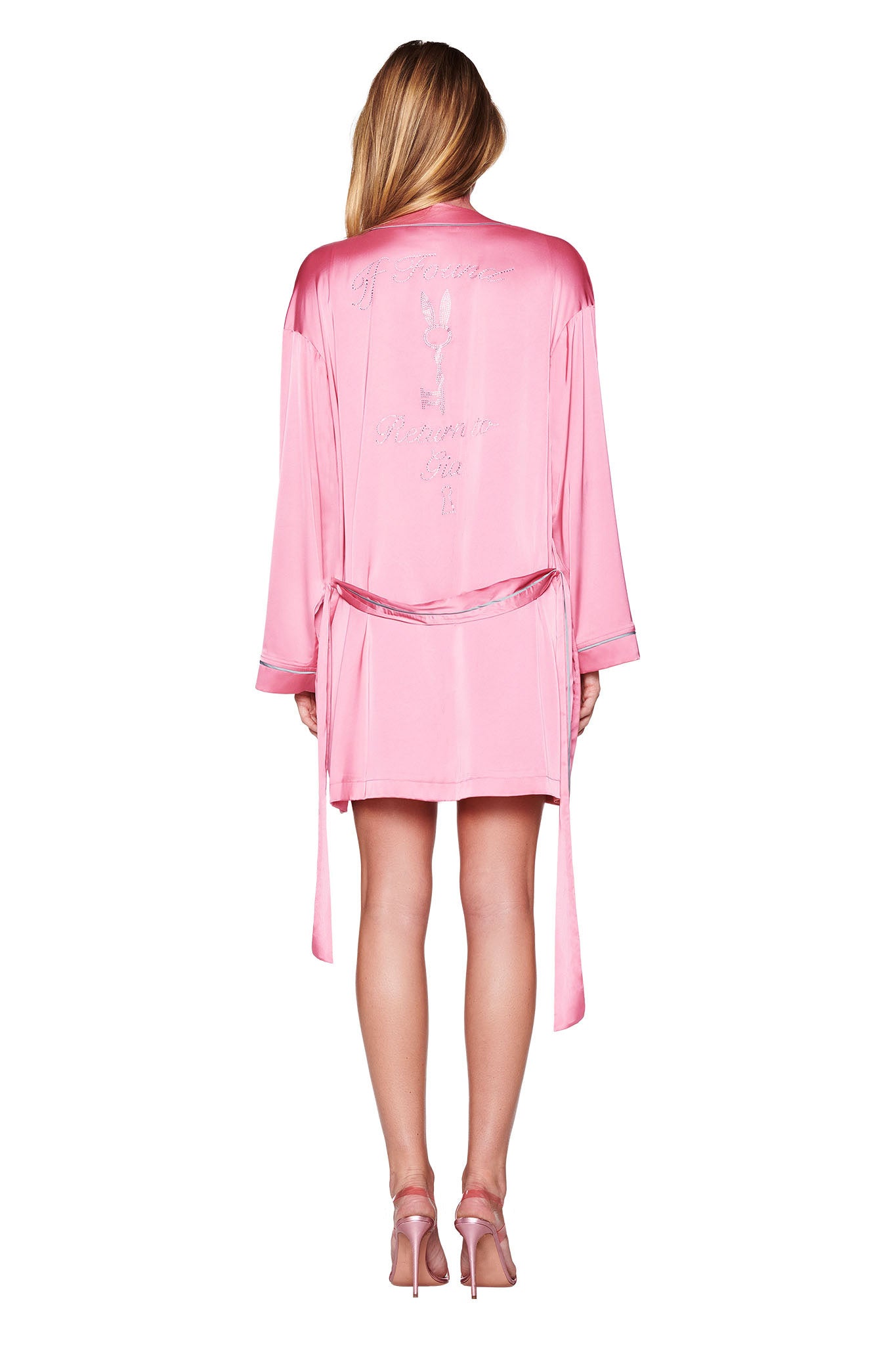 A person with long hair stands gracefully in a relaxed fit, the MARTINA SLEEP ROBE in pink, featuring custom I.AM.GIA diamante embroidery on the back. The satin robe, tied at the waist, pairs elegantly with high-heeled shoes against a plain white background.