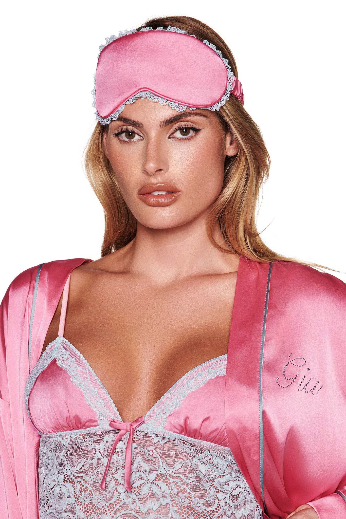 A person wearing the MARTINA SLEEP ROBE - PINK by I.AM.GIA, along with a coordinating pink sleep mask and lace-trimmed lingerie. The robe features a relaxed fit and 'GIA' stitched in cursive with custom diamante detailing. Their hair is down, and they maintain a neutral expression against a plain white background.