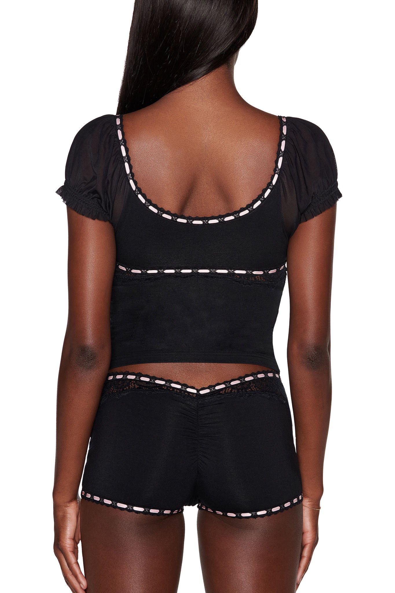 A person with long dark hair is seen from behind, wearing the DESTINY 2 PIECE SET - BLACK by I.AM.GIA. The outfit includes a black short-sleeve crop top and shorts featuring pink and white trim, with a bodycon fit, scoop neckline, and decorative lace trim details inspired by vintage sleepwear.