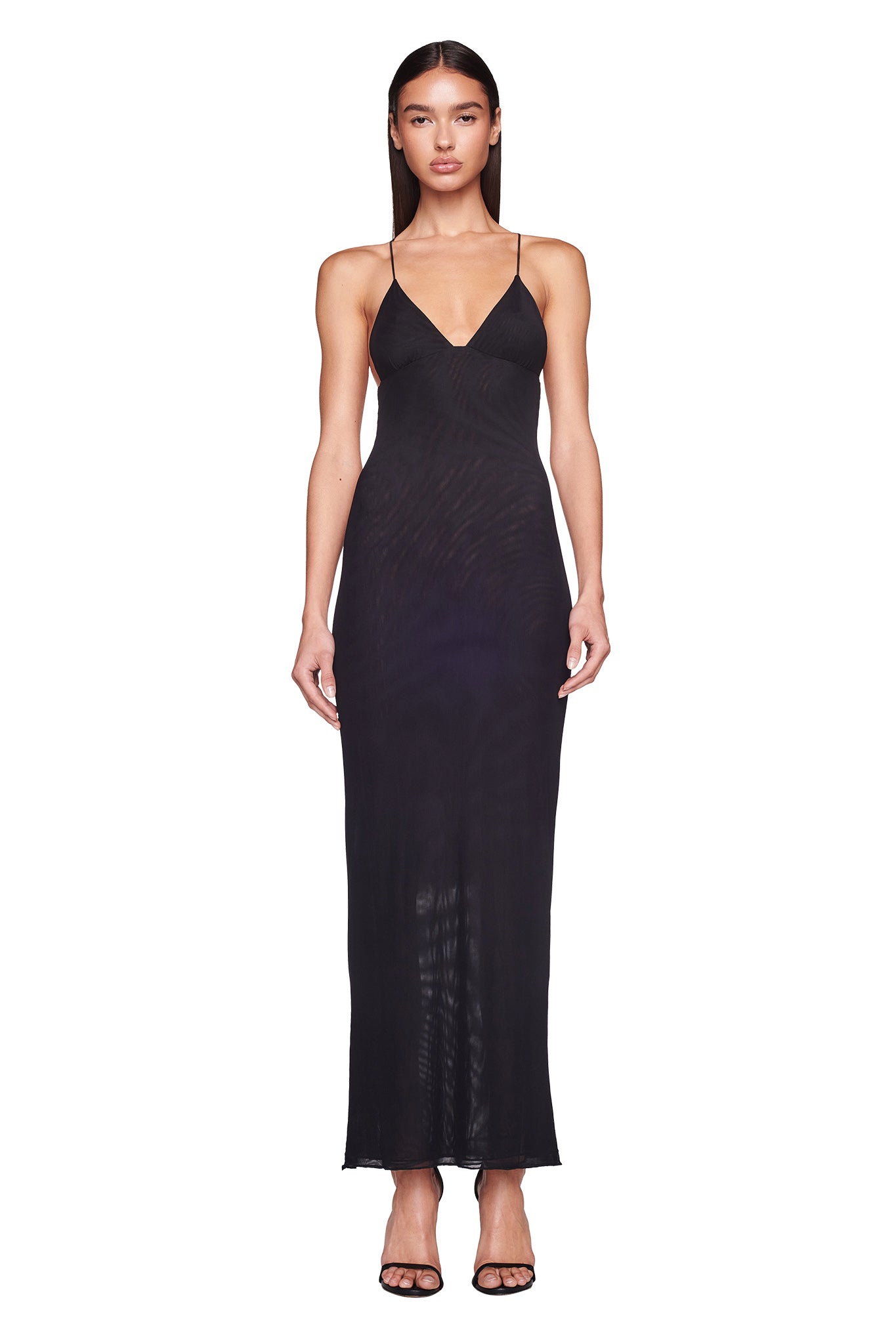 A woman stands against a plain white background, wearing the ELLERY DRESS - BLACK by I.AM.GIA, a long mesh maxi dress with spaghetti straps, an exposed open back, and a V-neckline. She has straight, long brown hair, minimal jewelry, and black high-heeled sandals.