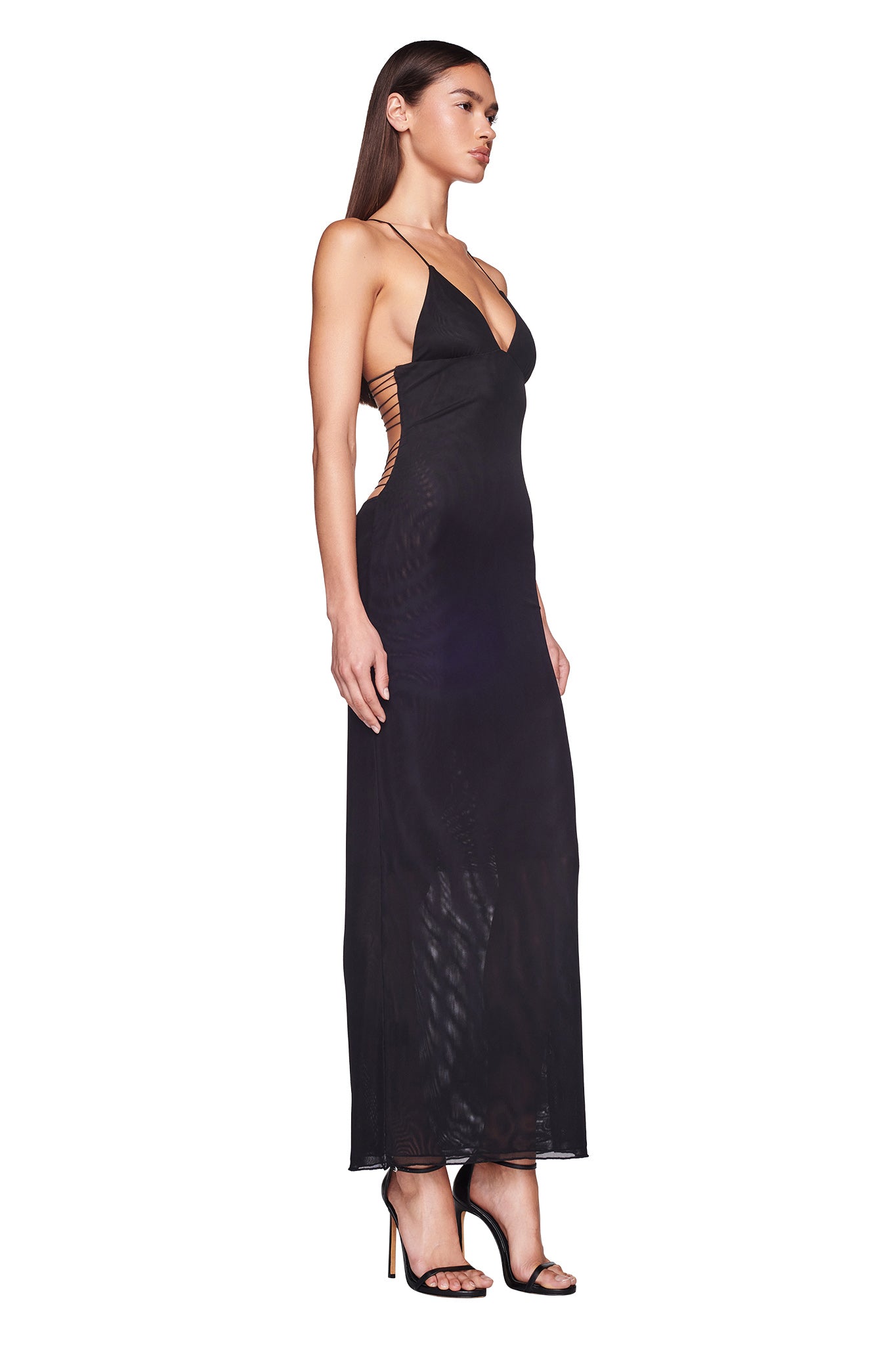 A woman stands in profile wearing the ELLERY DRESS - BLACK by I.AM.GIA. The long, elegant black mesh maxi dress features a deep V-neck and exposed open back detailing. With thin straps and a form-fitting design, she pairs the dress with black high-heeled sandals. Her straight, long hair complements her confident pose.