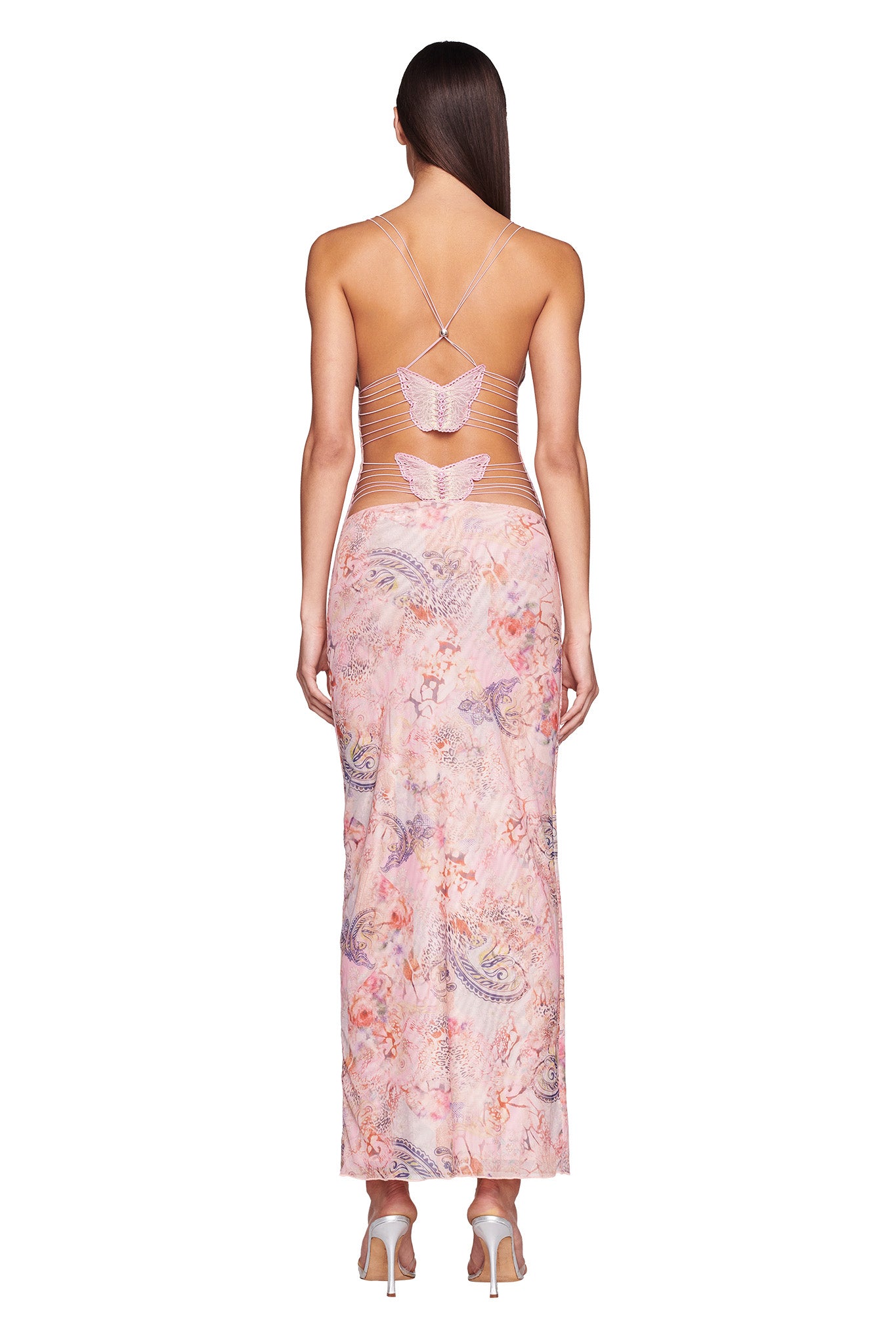 A woman with long brown hair is shown from the back, wearing the ELLERY DRESS - PINK : FLORA PAISLEY by I.AM.GIA, featuring a halter neck style with butterfly cut-out details on the back and an ankle-length floral pattern. She is standing in high-heeled shoes on a white background.