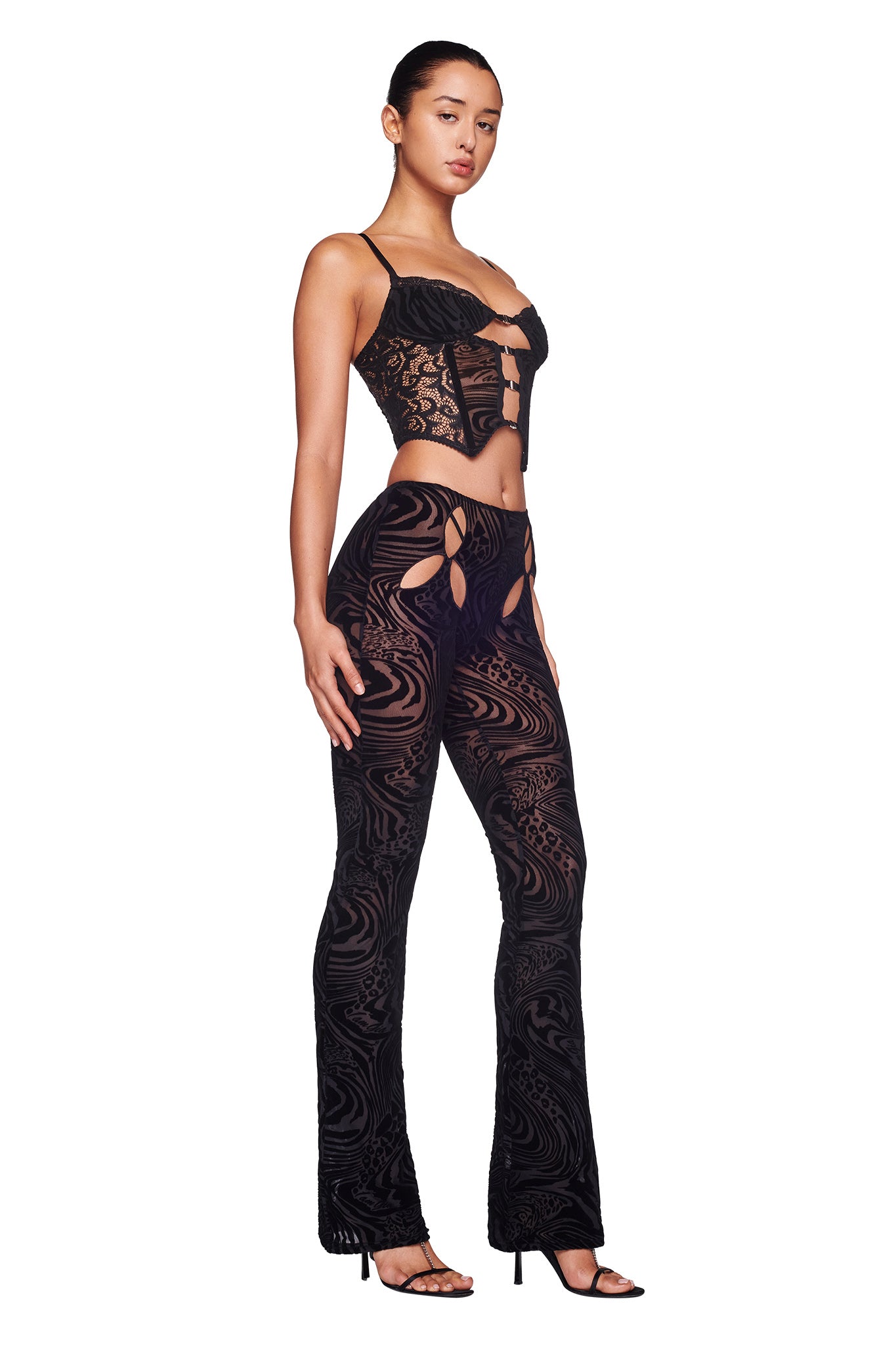A person poses confidently against a plain white background, wearing the ELENA TOP - BLACK BURNOUT by I.AM.GIA. The matching black lace outfit features a cropped top with thin straps and decorative cutouts, paired with high-waisted pants adorned with intricate patterns and cutouts.