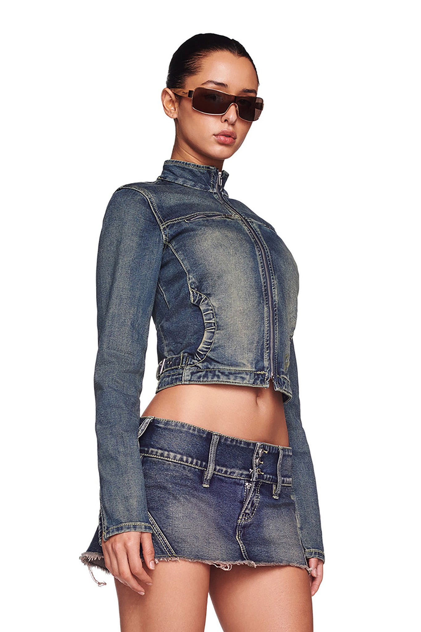 A person with long hair tied back wears large rectangular sunglasses and a fitted I.AM.GIA NYKKO JACKET - BLUE : DENIM BLUE with frayed edges at the hem, paired with a matching mini denim skirt. The outfit showcases a vintage wash and distressed look against a plain white background.