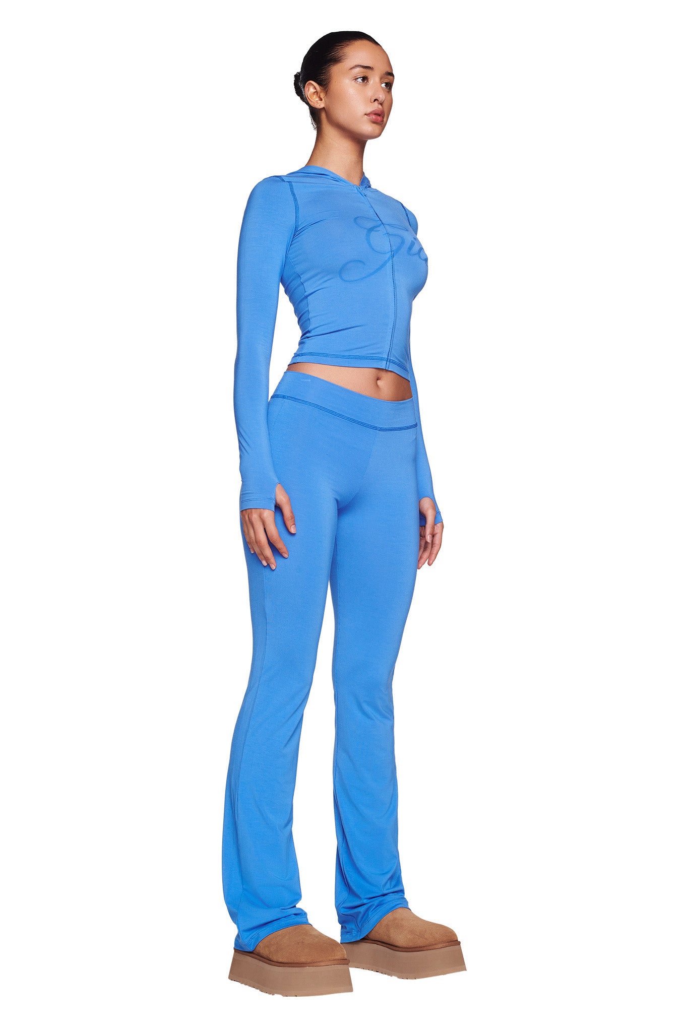 A person stands against a white background, wearing the I.AM.GIA BLARE TRACKPANT - BLUE set, paired with tan platform shoes. Their hair is neatly tied back. The outfit features a bodycon silhouette with thumb holes in the sleeves.