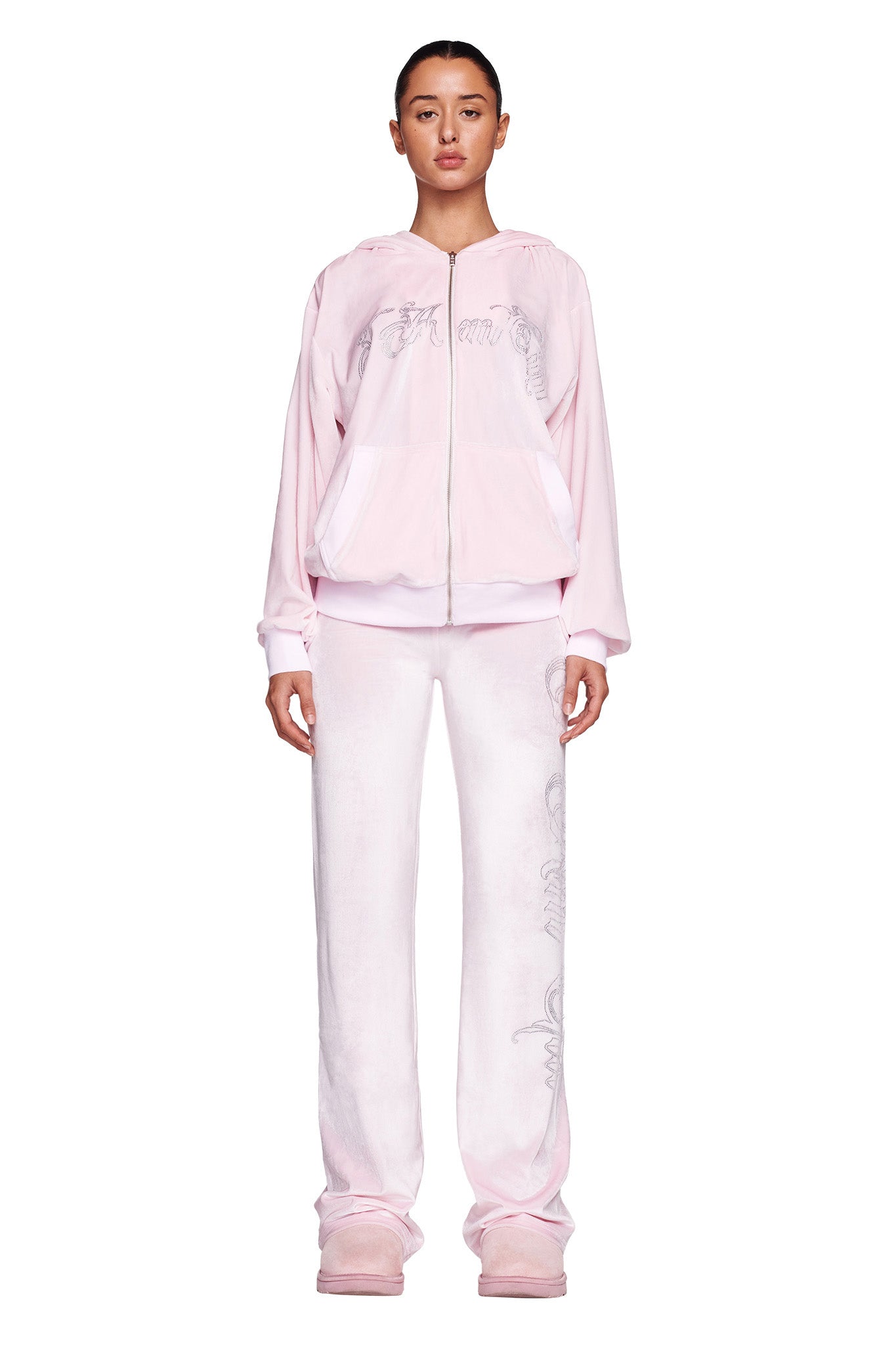 A person stands facing forward, sporting the I.AM.GIA Daria Hoodie in icy pink velvet with a hooded zip-up jacket and matching pants. The outfit features subtle text details. They are also wearing light pink shoes against a plain white background.