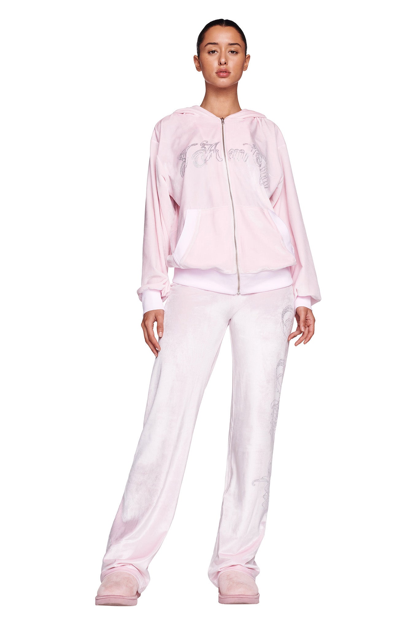 A person stands facing forward, dressed in an icy pink velour tracksuit from I.AM.GIA, featuring a zip-up jacket and matching DARIA PANT - PINK : ICY PINK. The outfit is adorned with subtle embroidered details and the custom 'I.Am.GIA' logo. The individual is also wearing light pink slippers against a white background.